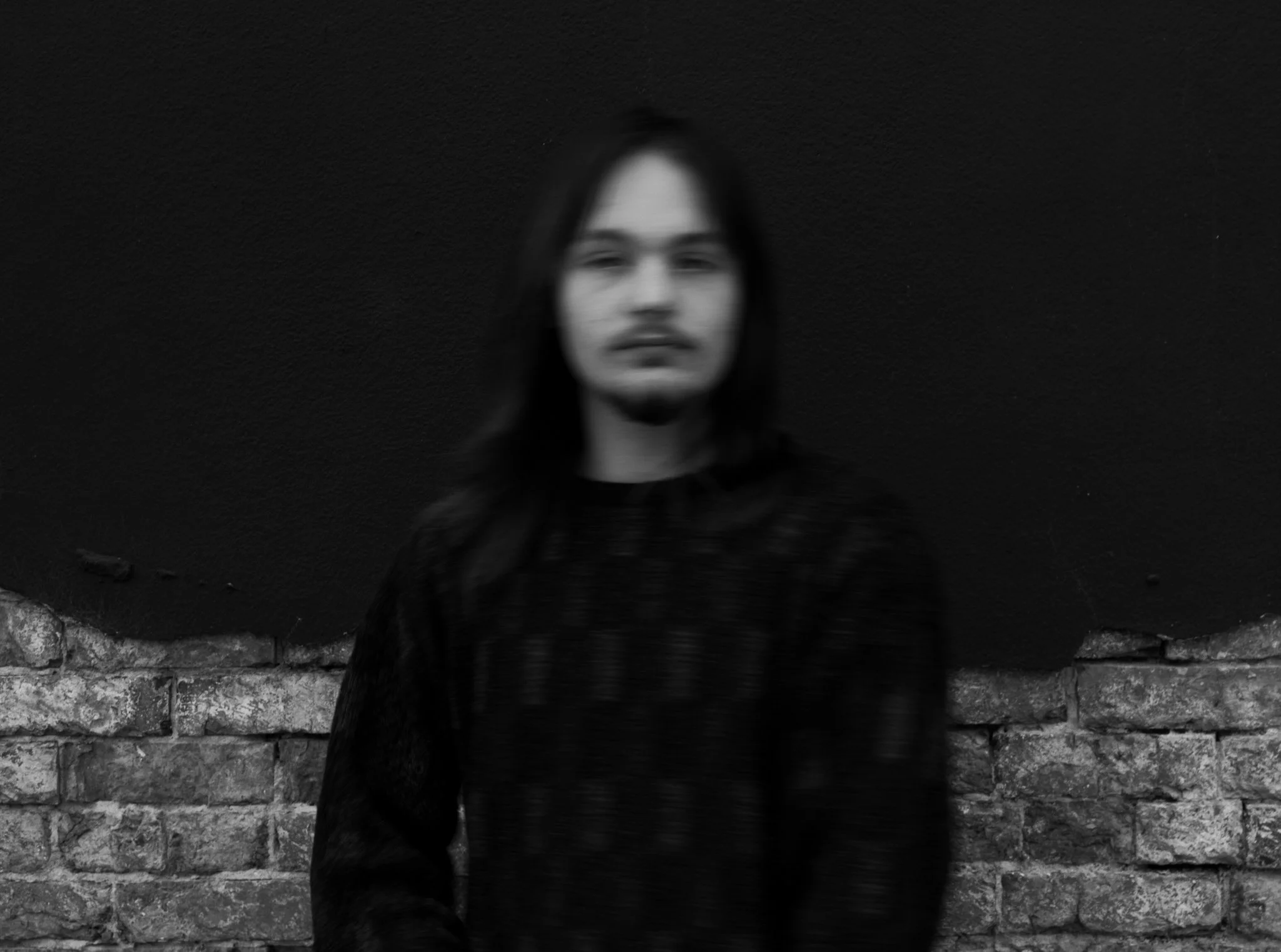 A person with long hair and a slight beard standing in front of a brick wall and black background, appearing blurred.