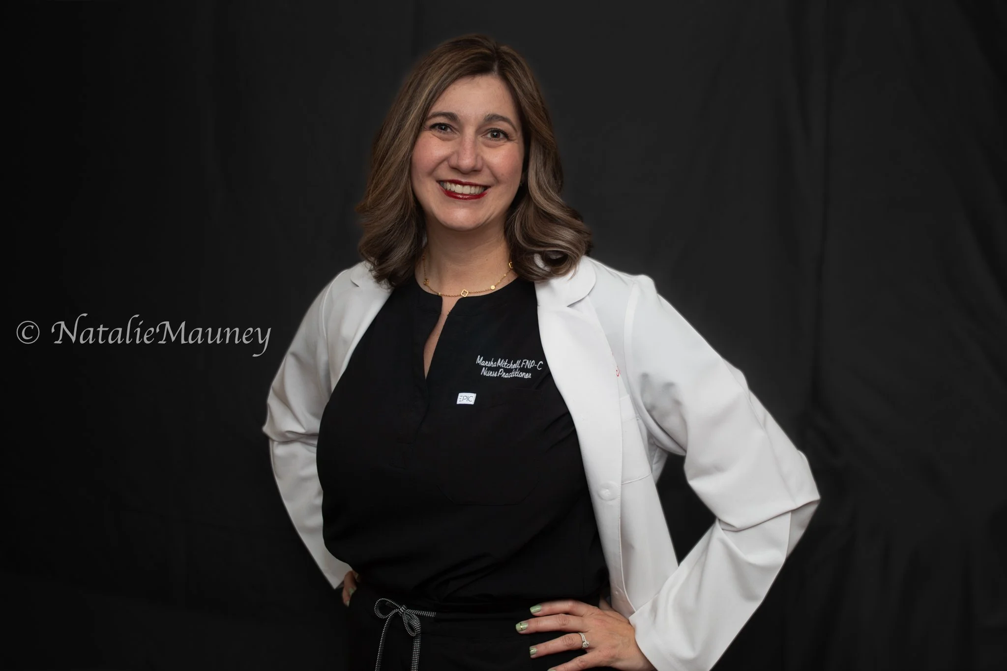 A woman with brown wavy hair, wearing black scrubs and a white lab coat, smiling and standing with hands on hips against a black background.