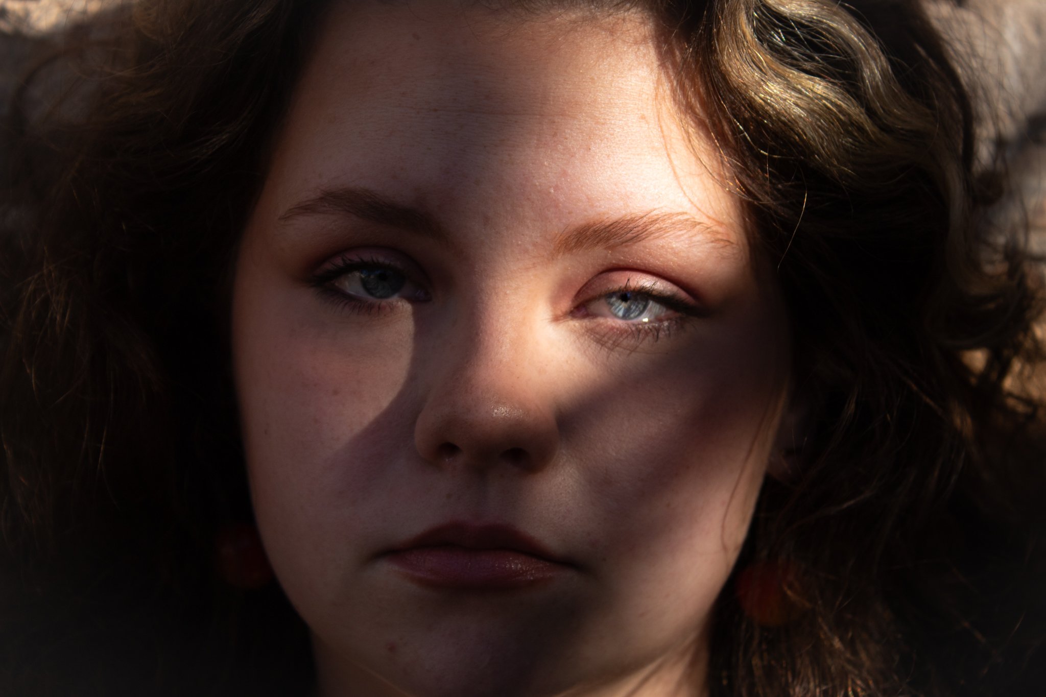 Close-up photo of a young woman's face with one eye in shadow and the other illuminated by sunlight, revealing her blue eye and brunette hair.
