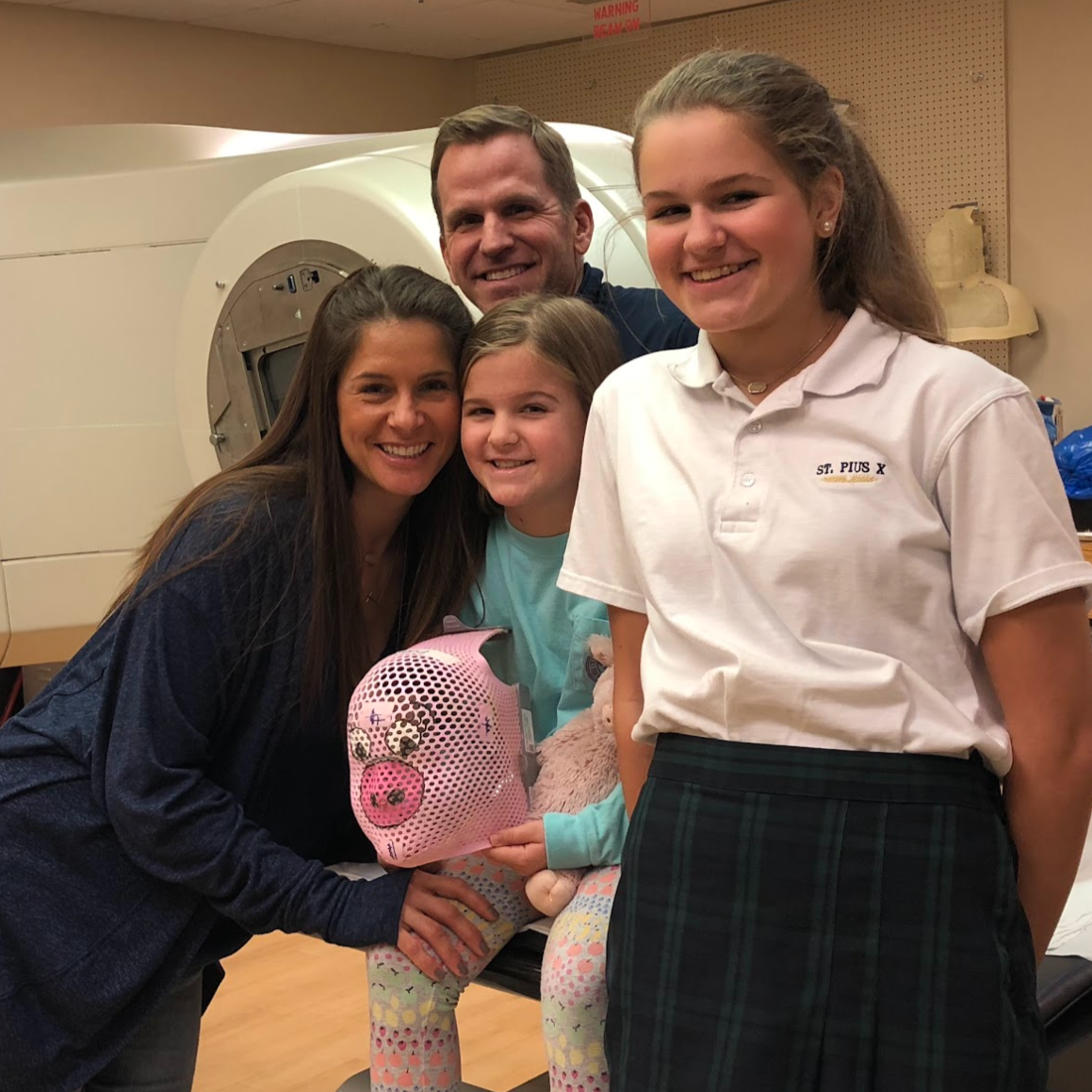 A group of five people smiling in a medical setting, with a girl holding a pig-shaped piggy bank, standing in front of a hospital MRI machine.