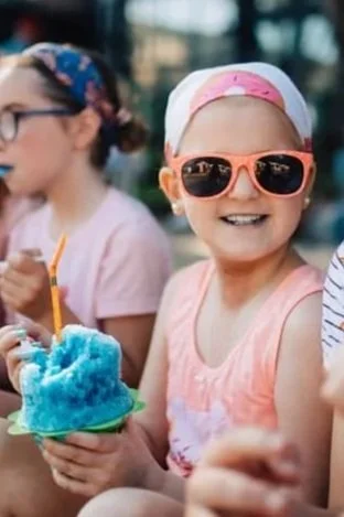 A young girl wearing orange sunglasses, a white cap with pink accents, and a pink sleeveless top, smiling and holding a blue cotton candy. Other children are partially visible in the background.