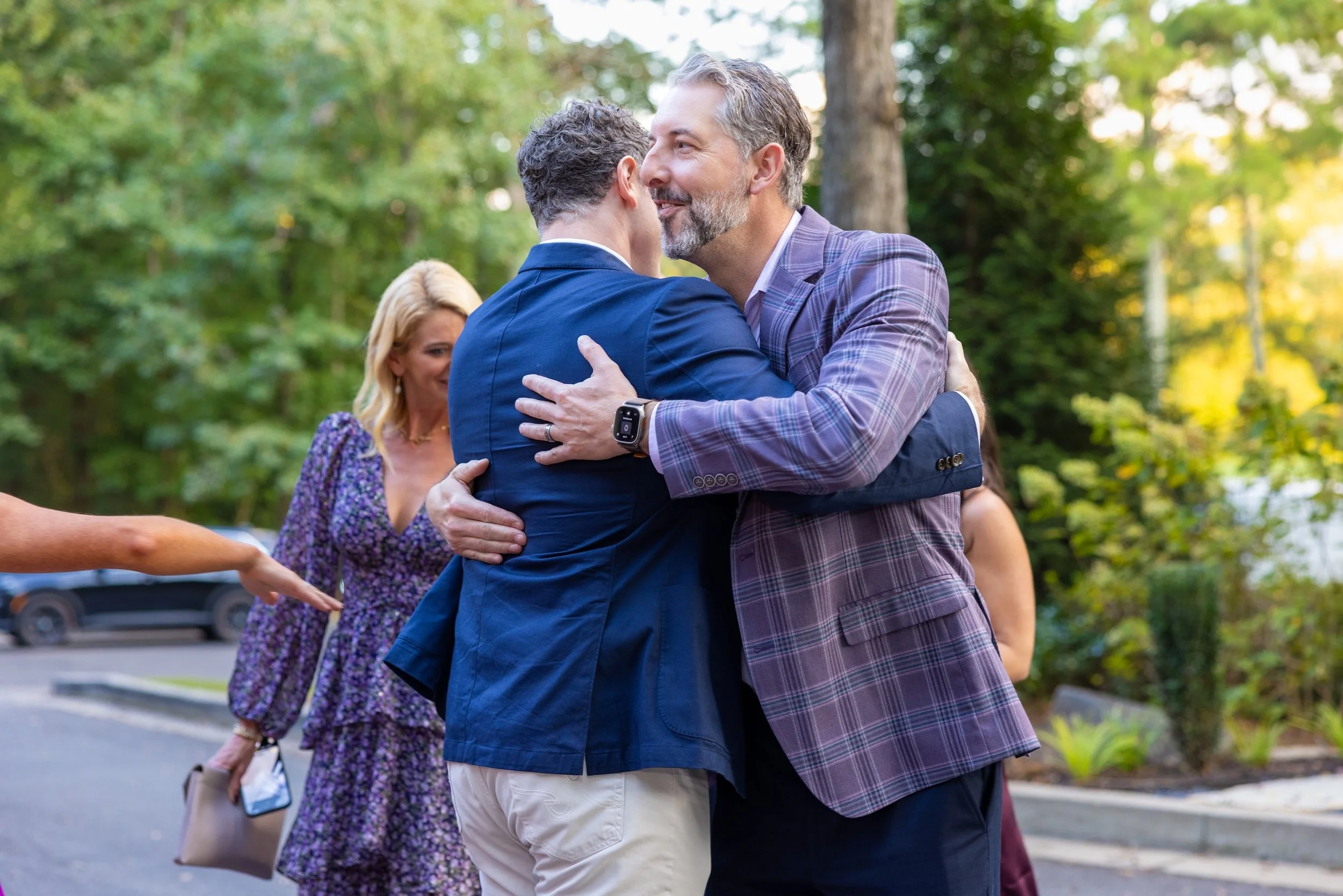 Two men in suits hugging each other outdoors in a park, with a woman in a purple dress standing nearby. The background features green trees and sunlight.