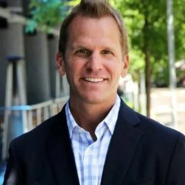 A smiling man with short hair wearing a dark suit jacket and a light blue checkered shirt, standing outdoors with trees and buildings in the background.