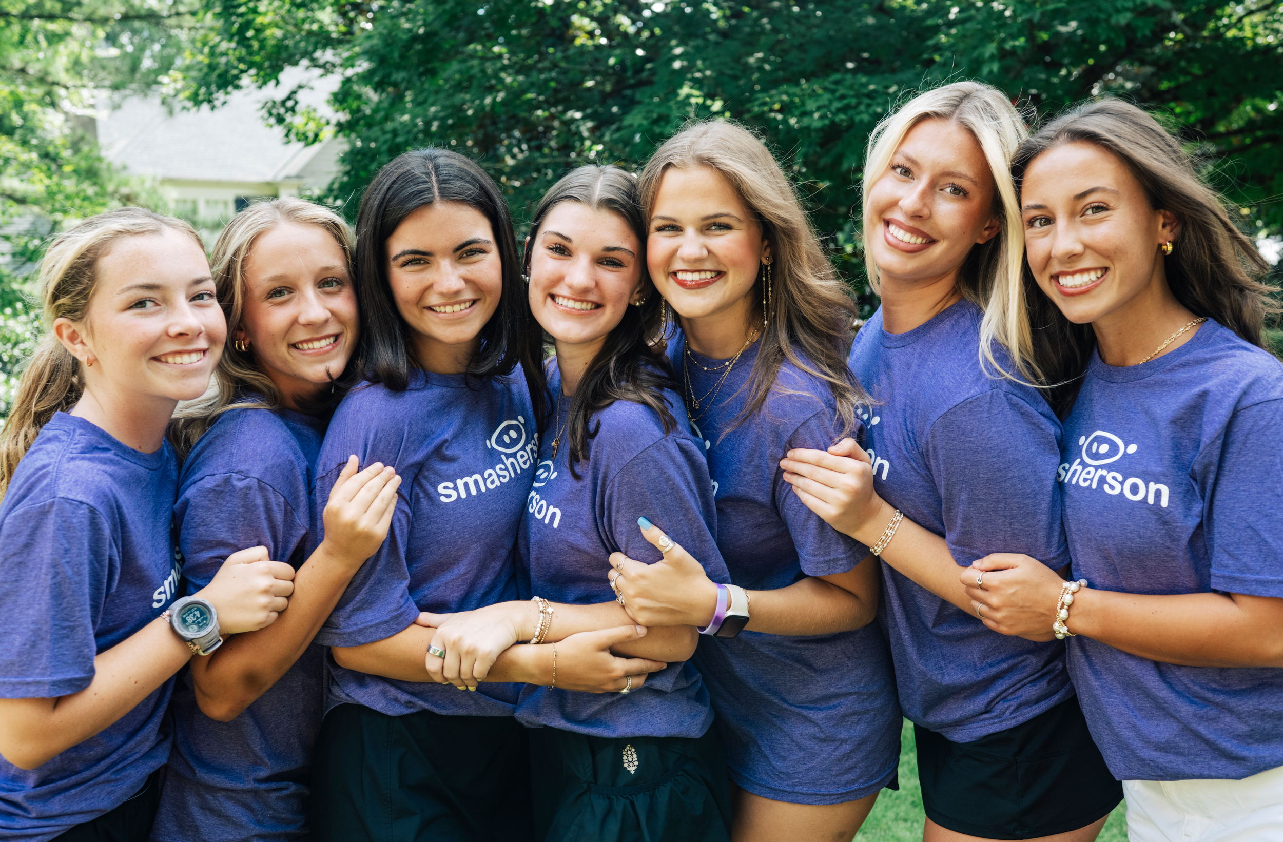 A group of seven young women smiling and hugging outdoors, wearing matching blue T-shirts with a logo that reads 'smashers'.