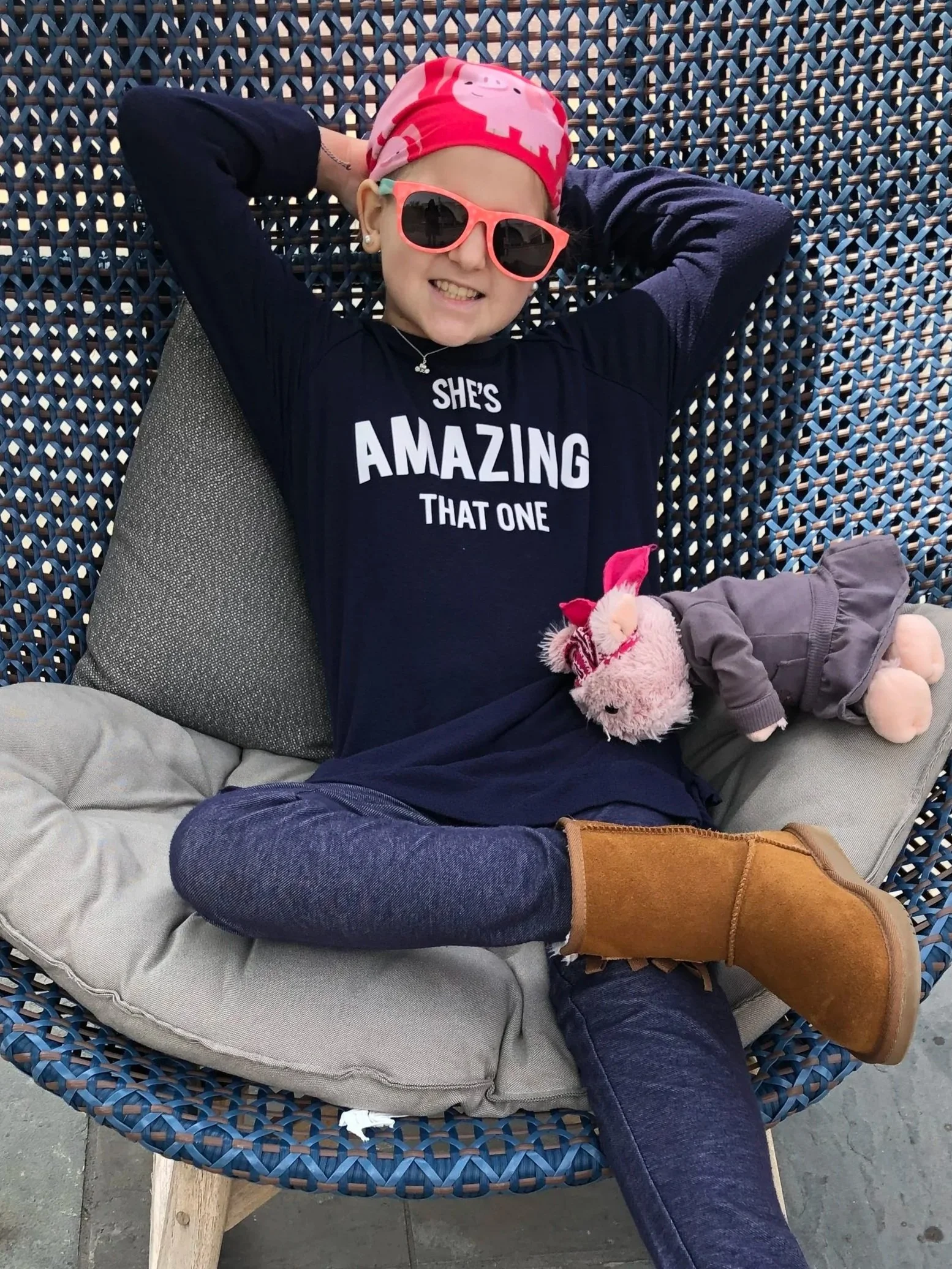 A girl with a pink and white piggy bank headband, pink sunglasses, a black shirt with white text, and jeans, sitting on a woven outdoor chair with a gray cushion, smiling and relaxing with her arms behind her head. A stuffed animal is on her lap.