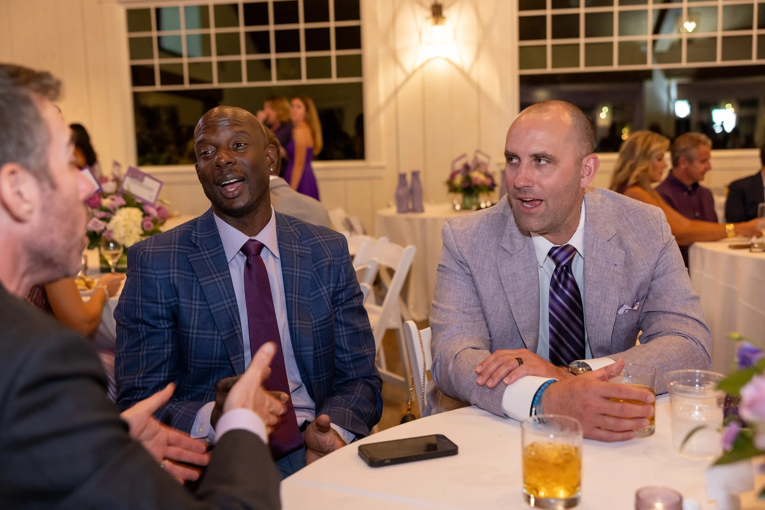 Four men in formal attire engaged in conversation at a dinner party, with drinks and floral centerpieces on the table, in a well-lit banquet hall with large windows.