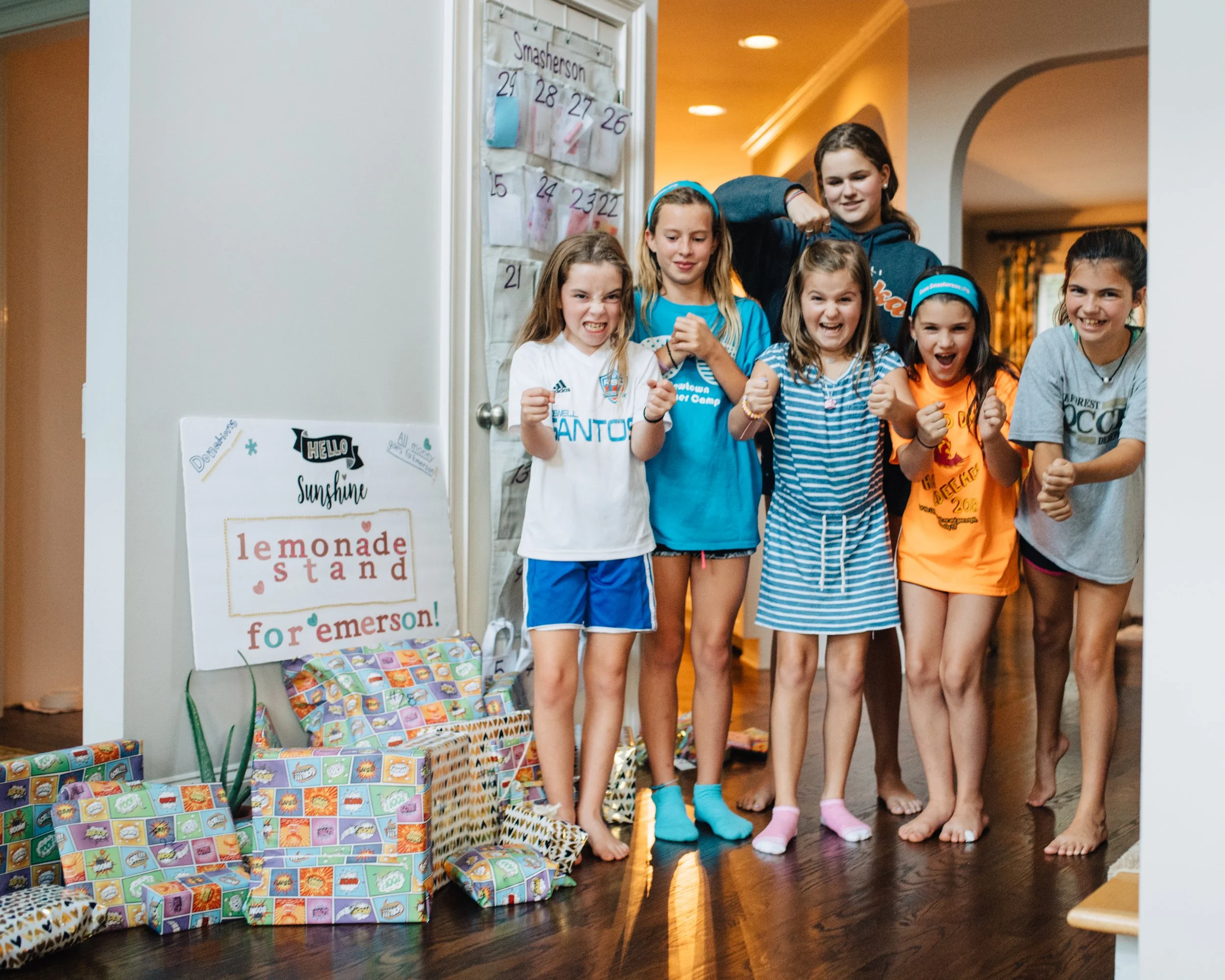 Group of children indoors celebrating, standing in front of a wall with a calendar and colorful wrapped presents on the floor.