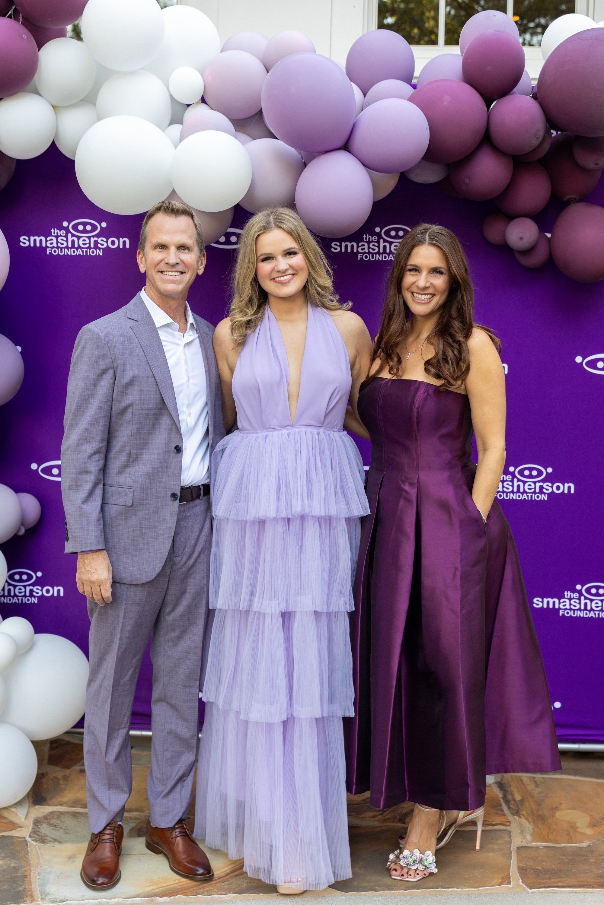 Three people standing in front of a purple backdrop with balloons and the logo of The Smasher Foundation. The man on the left is wearing a light gray suit and brown shoes. The woman in the middle is wearing a lavender dress with ruffles. The woman on