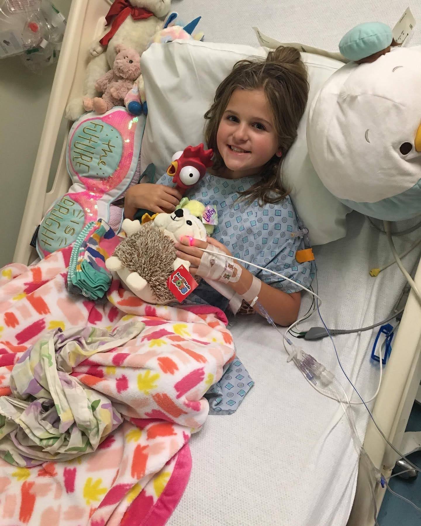 Child lying in a hospital bed with various stuffed animals and toys around her, holding some plush toys, with medical equipment and IV line attached.