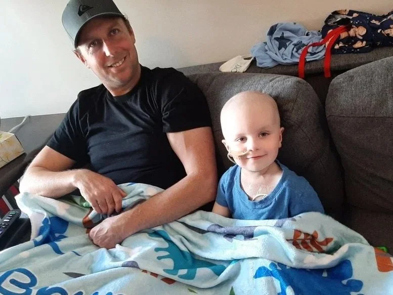 A man and a young boy sitting on a couch. The man is smiling and wearing a gray cap and black shirt. The boy has a medical tube in his nose, is dressed in a blue shirt, and is covered with a colorful blanket. There are clothes and a red item on the couch behind them.