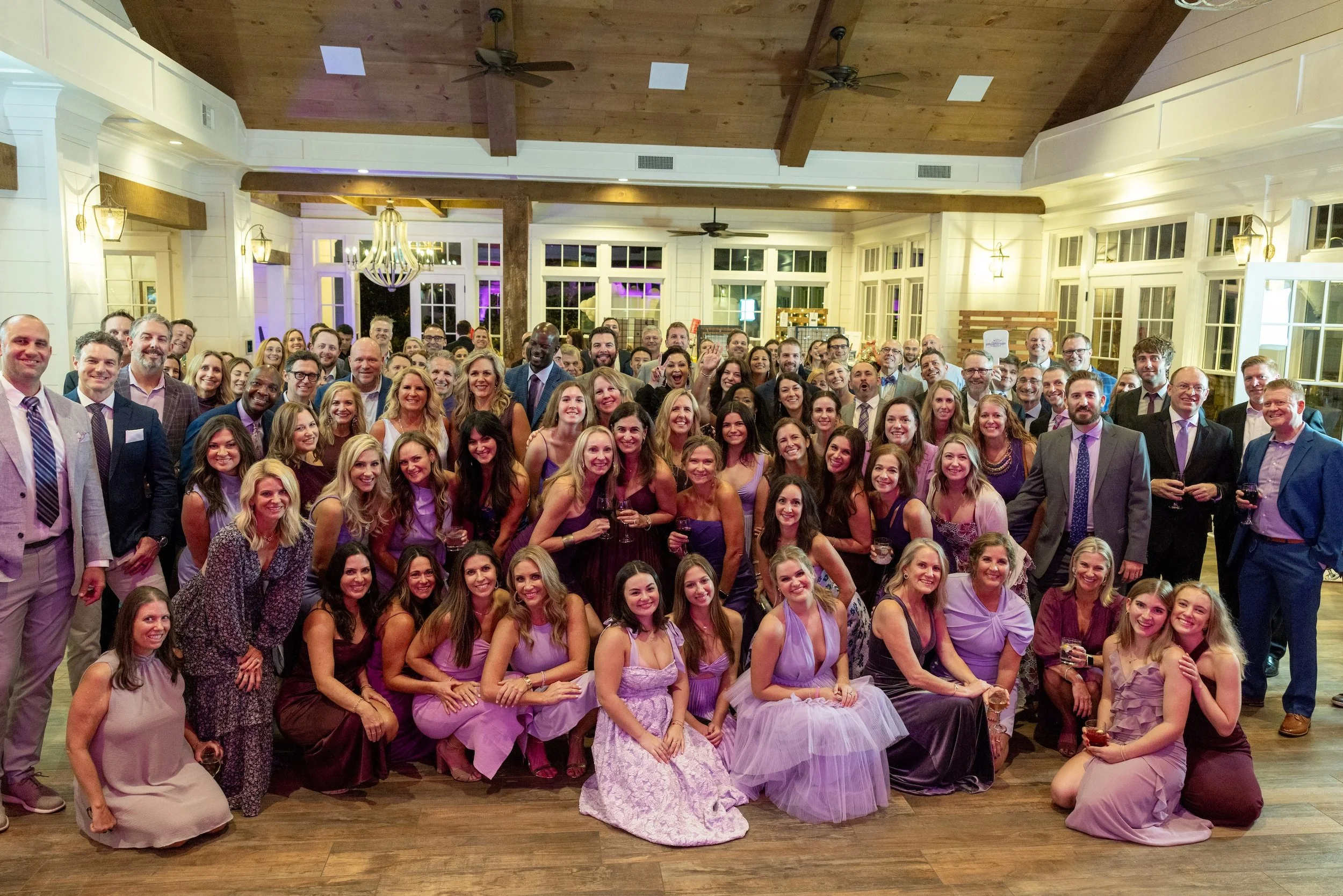 A large group of people dressed in formal attire gathered together indoors for a celebration or event, smiling and posing for a group photo under wooden ceilings with ceiling fans and large windows.