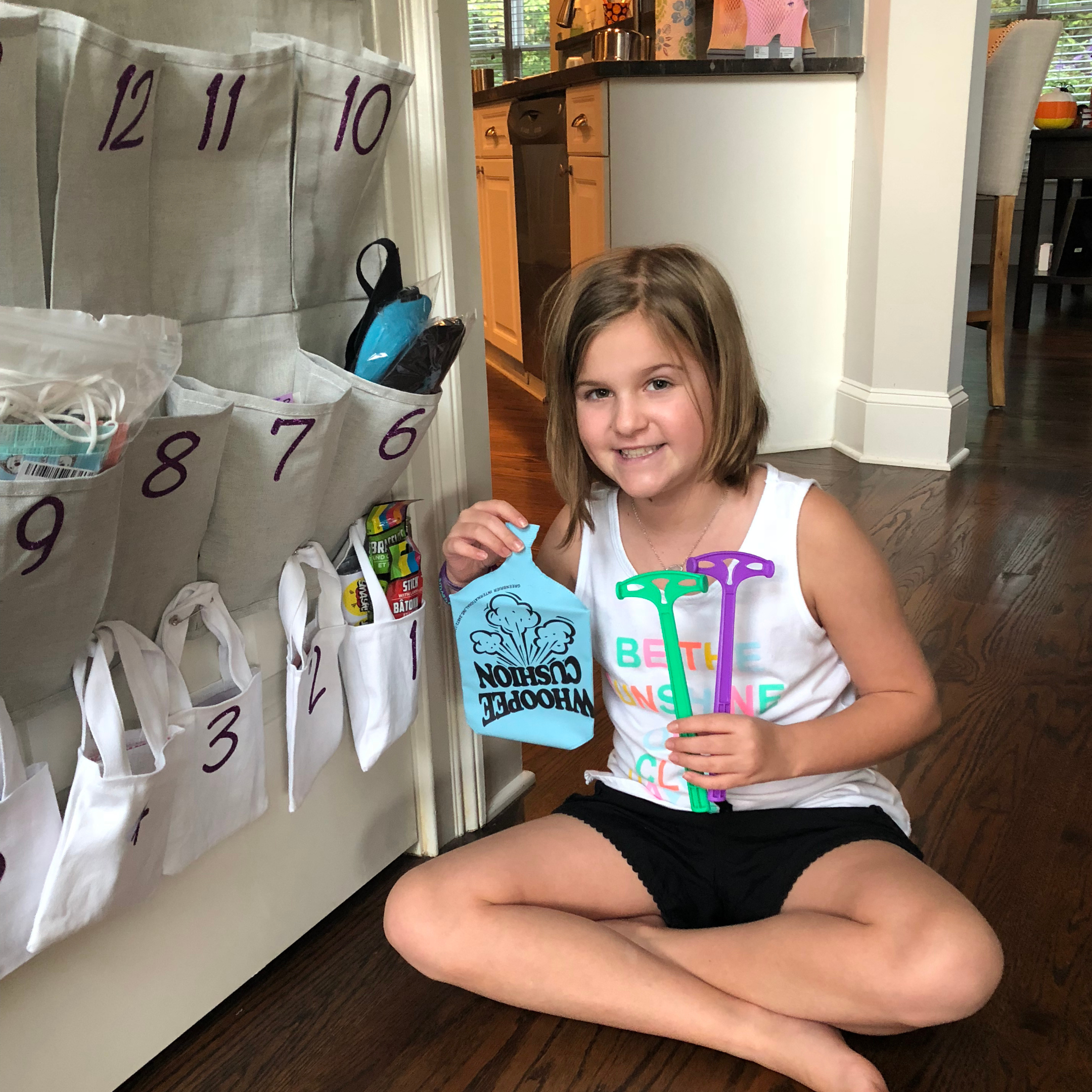A young girl with shoulder-length brown hair smiling while holding colorful birthday goodie bags, sitting on the floor near a row of fabric bags with purple handwriting numbered 6 to 12 hanging on a wall in a room with wooden flooring, white walls, and visible kitchen and dining area in the background.