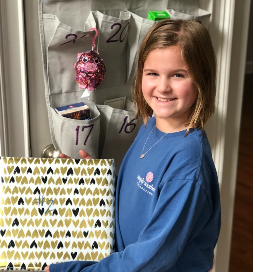 A young girl with shoulder-length light brown hair smiling, wearing a blue long-sleeve shirt with a school logo, standing next to a calendar with a gold and black heart pattern.
