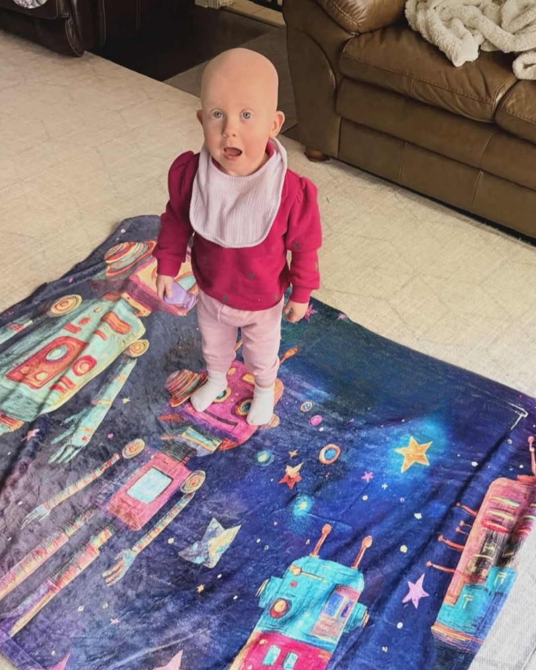 A baby with no hair, wearing a pink long-sleeve shirt, white bib, light pink pants, and white socks, standing on a colorful space-themed blanket with rockets and stars, on a beige carpet in a living room with a brown couch and blanket in the background.