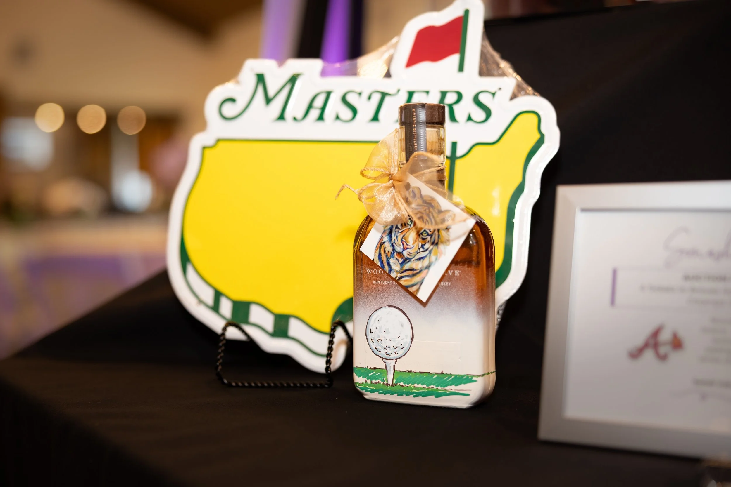 Close-up of a decorative sign featuring the Kentucky Bourbon Trail logo, with a small bottle of whiskey and a tag showing a tiger illustration hanging in front of it.