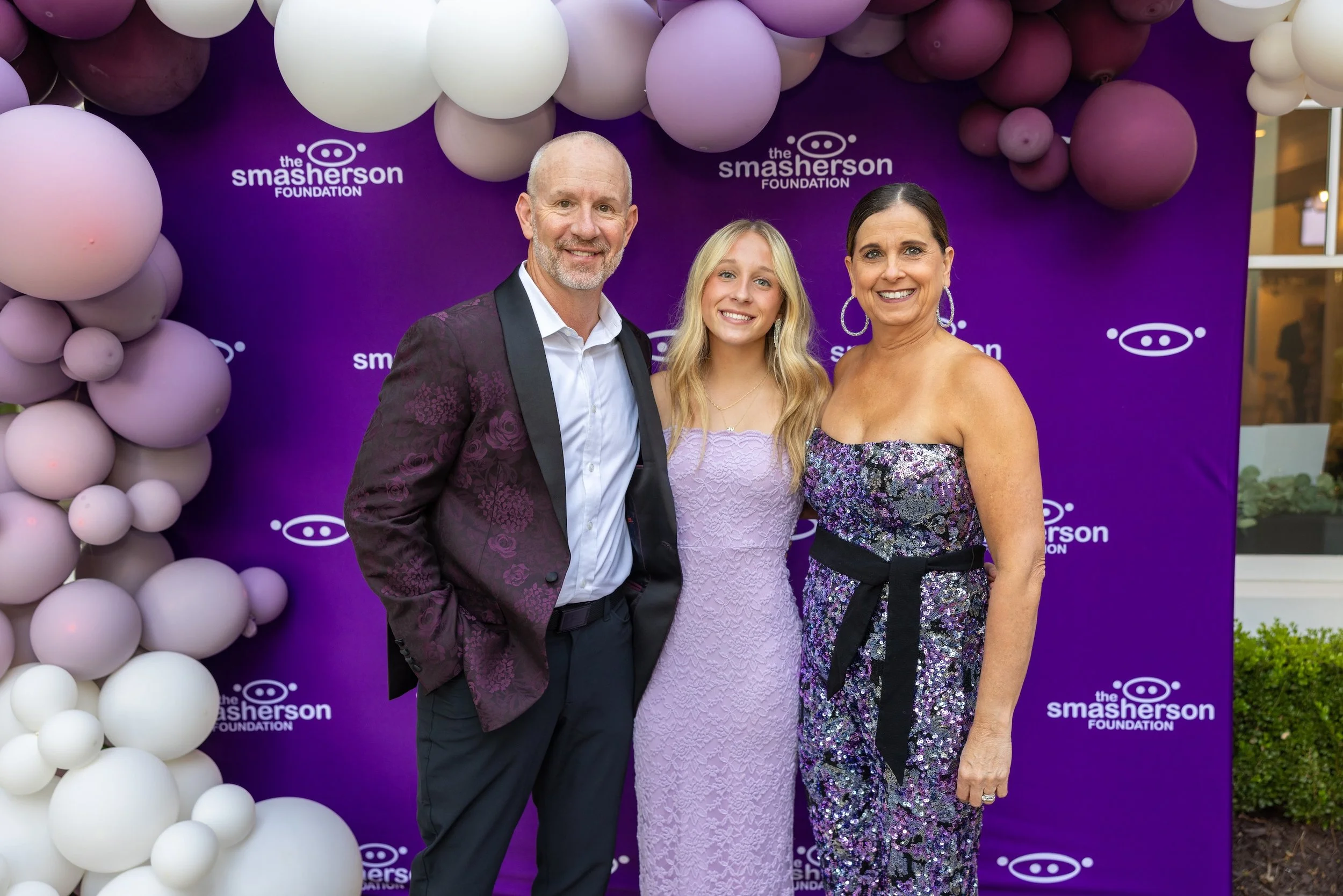 Three people standing in front of a purple backdrop with the logo 'the Smasherson Foundation' and white balloons. The man on the left is bald with a gray beard, wearing a dark patterned suit and white shirt. The woman in the middle has blonde hair, w