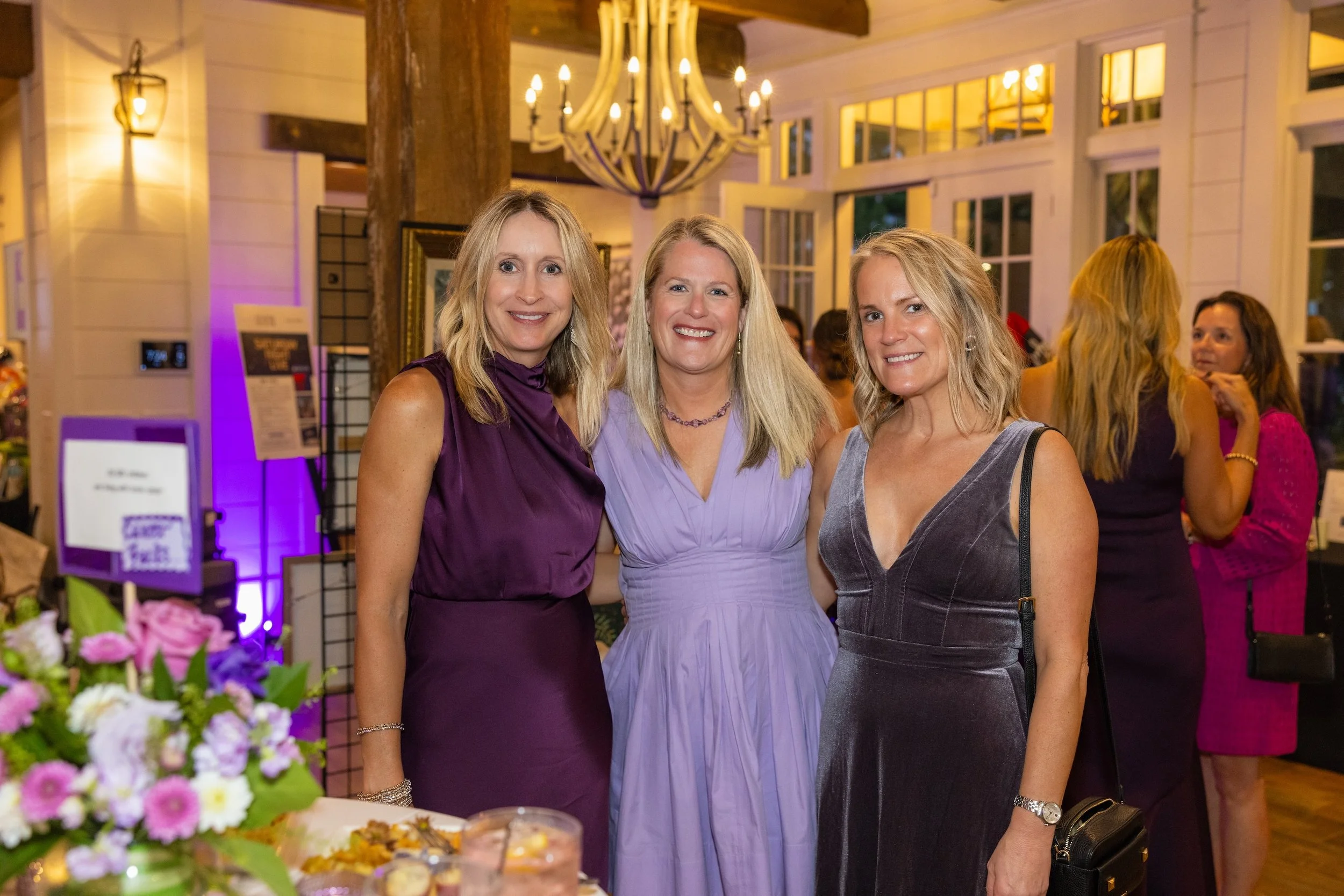 Three women standing together at an indoor event, smiling at the camera. They are dressed in elegant dresses, with flowers and a dessert table in front of them, and a chandelier hanging from the ceiling behind them.