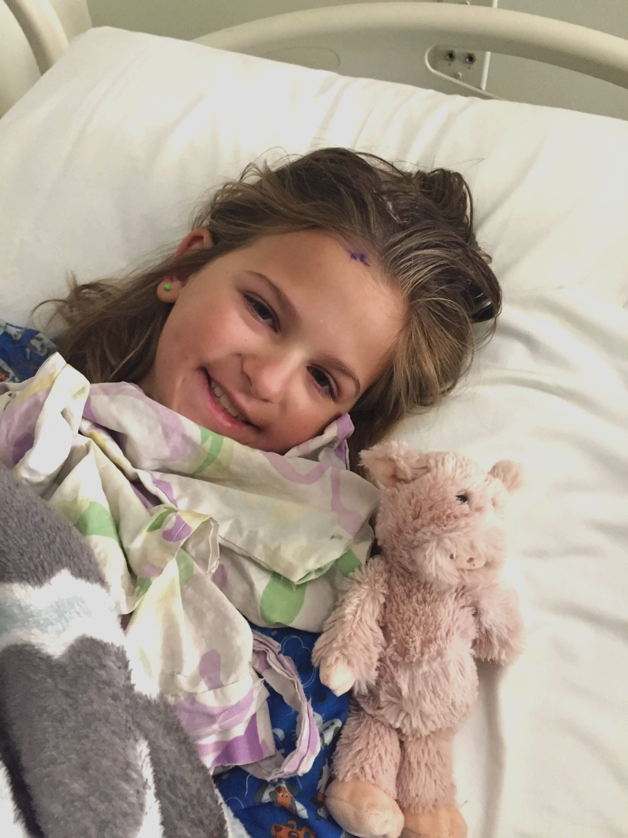 A young girl lying in a hospital bed smiling, holding a pink teddy bear, wearing blue pajamas with cartoon characters, and a hospital gown with pastel colors.