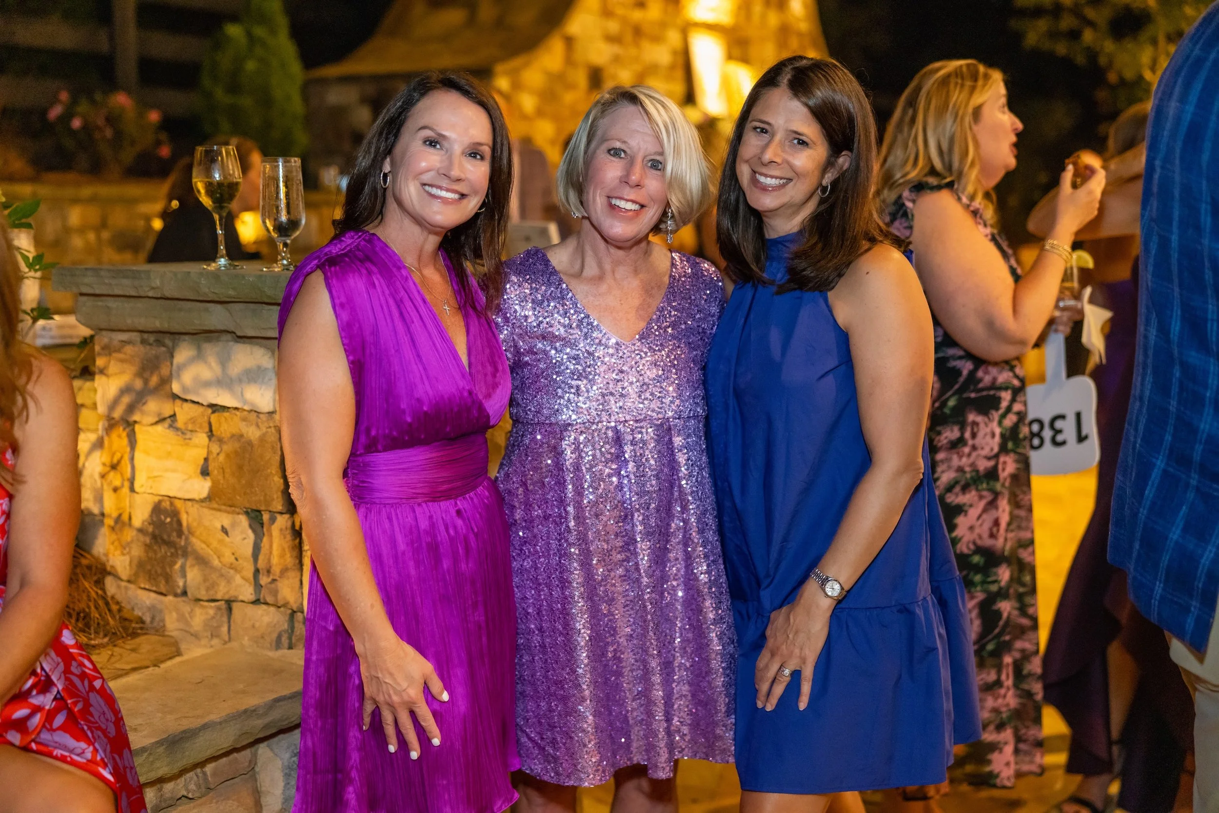 Three women standing together smiling at a nighttime outdoor event. They are dressed in purple, lavender, and blue dresses. There are glasses of wine or champagne on a stone ledge nearby, with other people in the background.