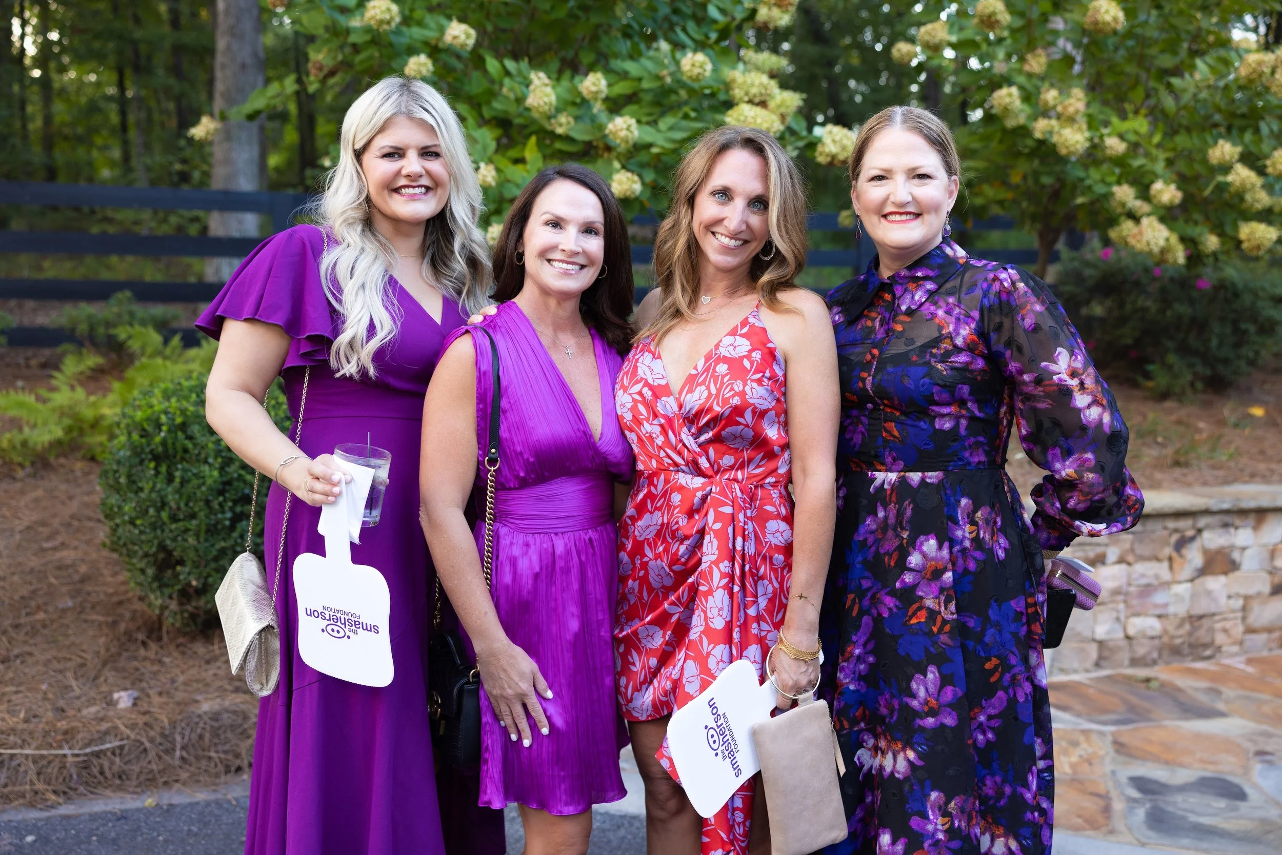 Four women dressed in purple, red, and floral dresses smiling outdoors in front of trees and bushes, holding program or event signage.