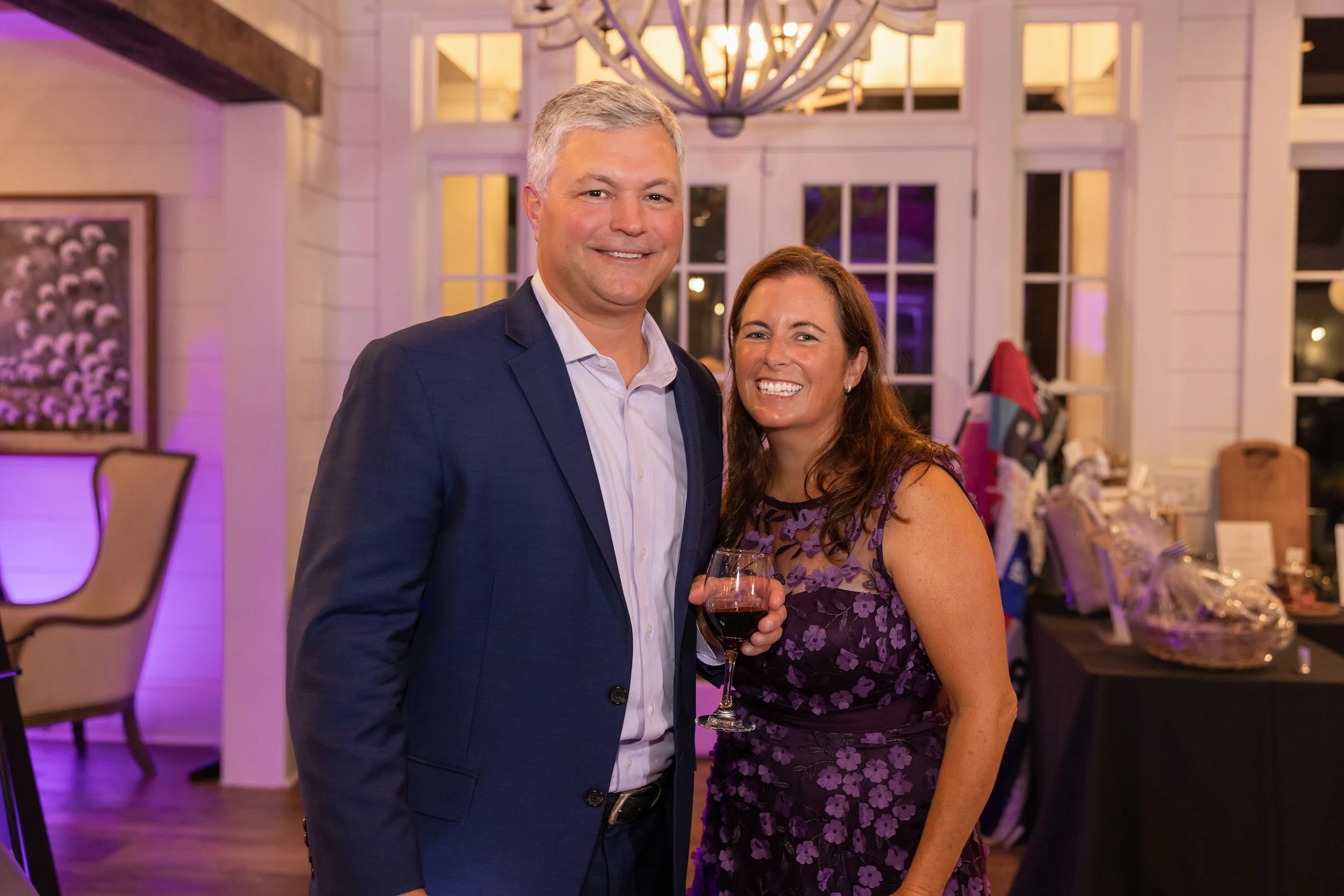 A man in a navy suit and a woman in a purple floral dress smiling at the camera at an indoor event, holding a glass of red wine, with purple lighting and decorations in the background.