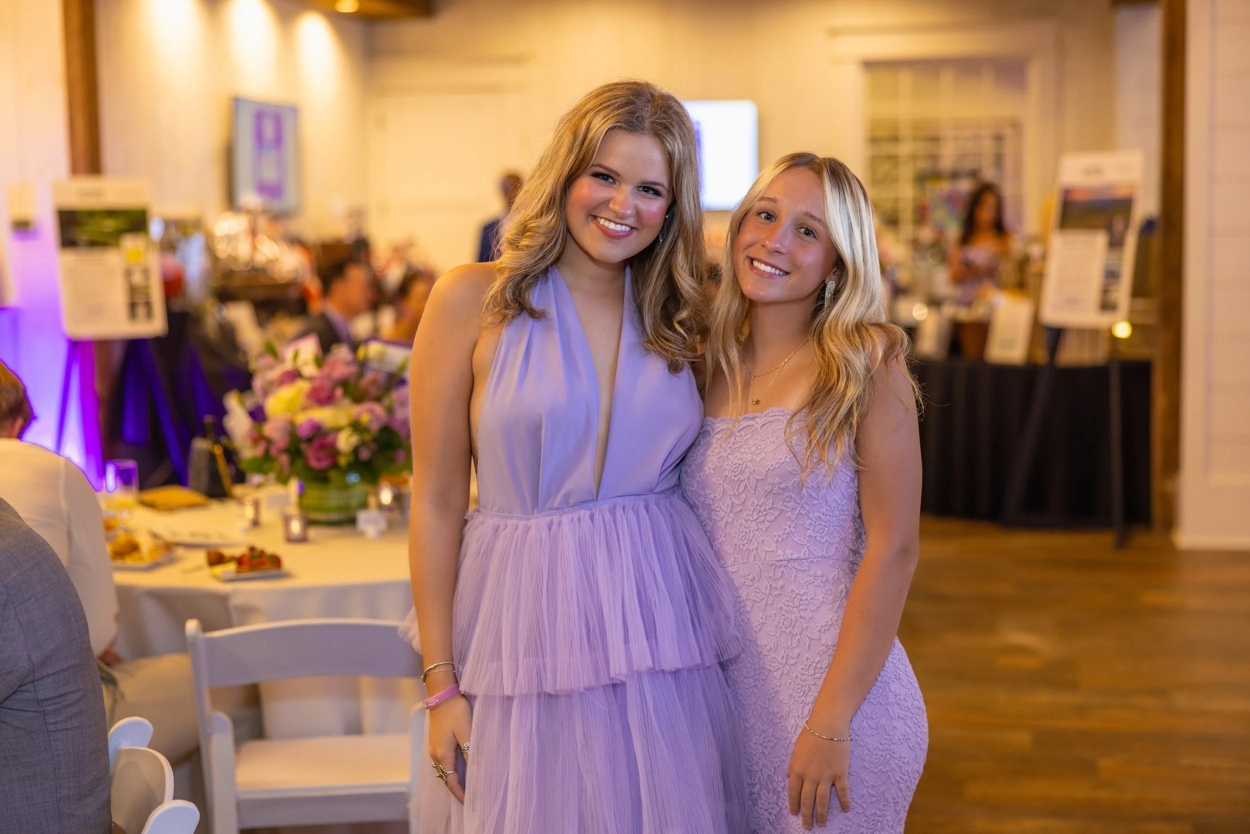 Two young women with blonde hair smiling at the camera, dressed in light purple dresses, at a social event with tables and flower arrangements in the background.