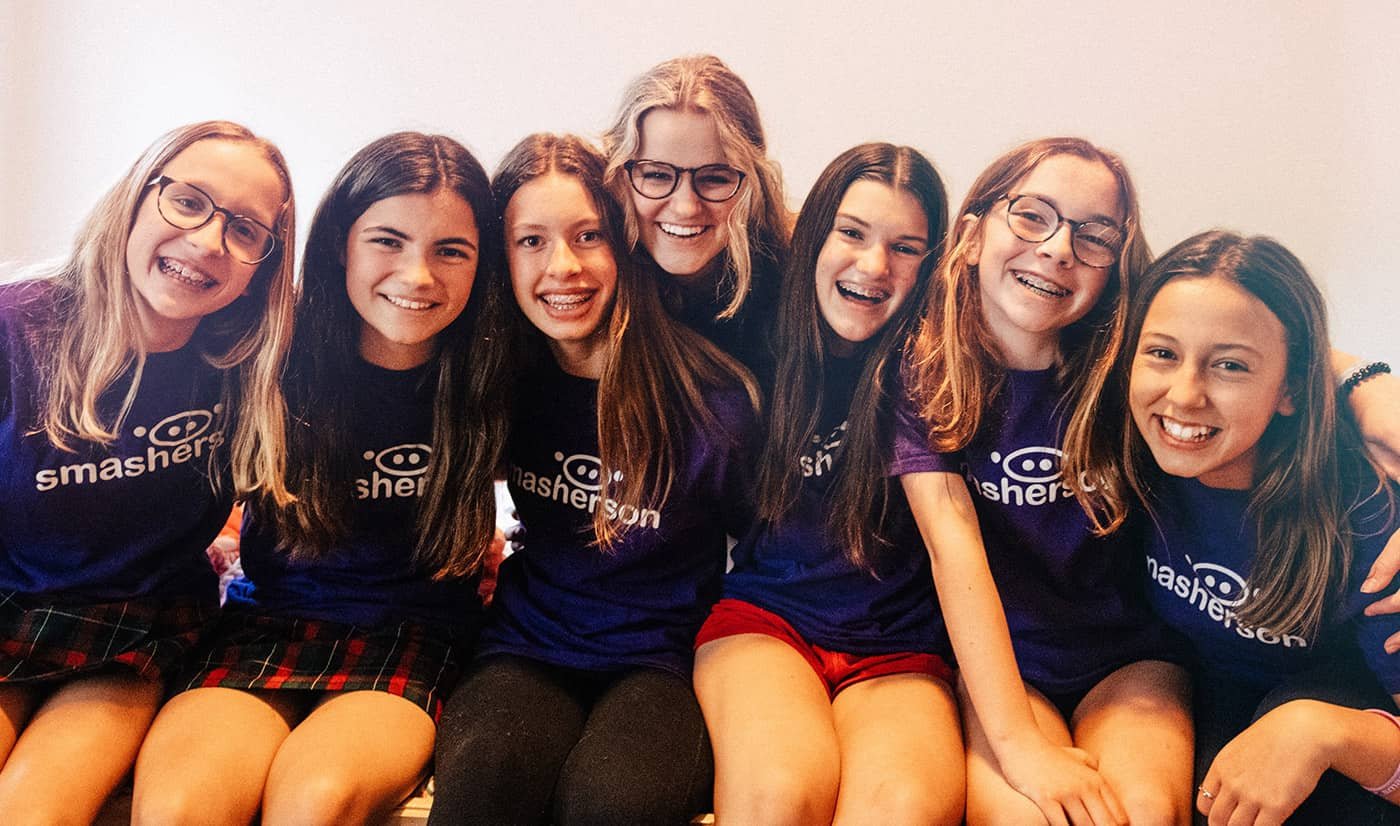 Group of seven smiling young girls sitting together, wearing matching navy blue T-shirts with 'smasher' logo, and casual clothing.