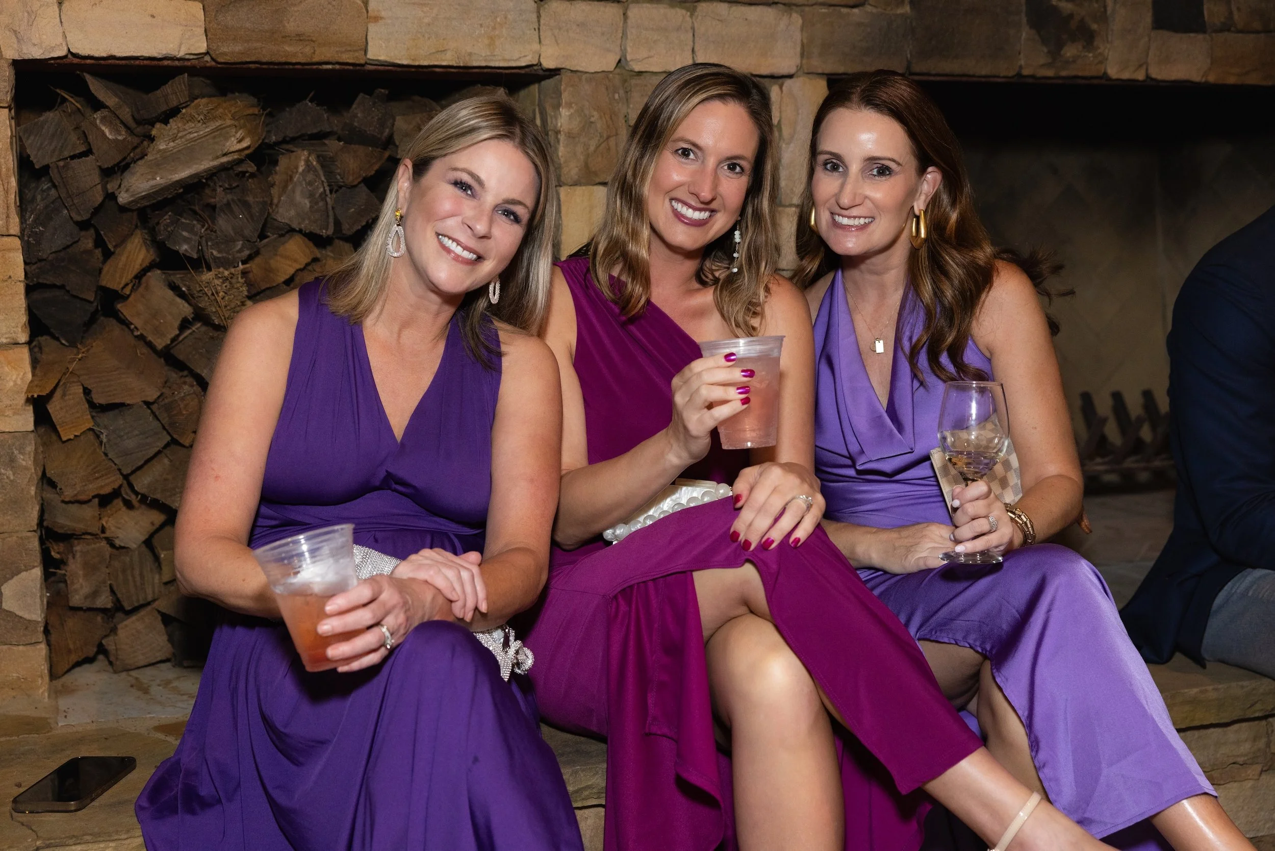 Three women sitting together smiling at a social gathering, dressed in purple and pink dresses, holding drinks near a stone fireplace.