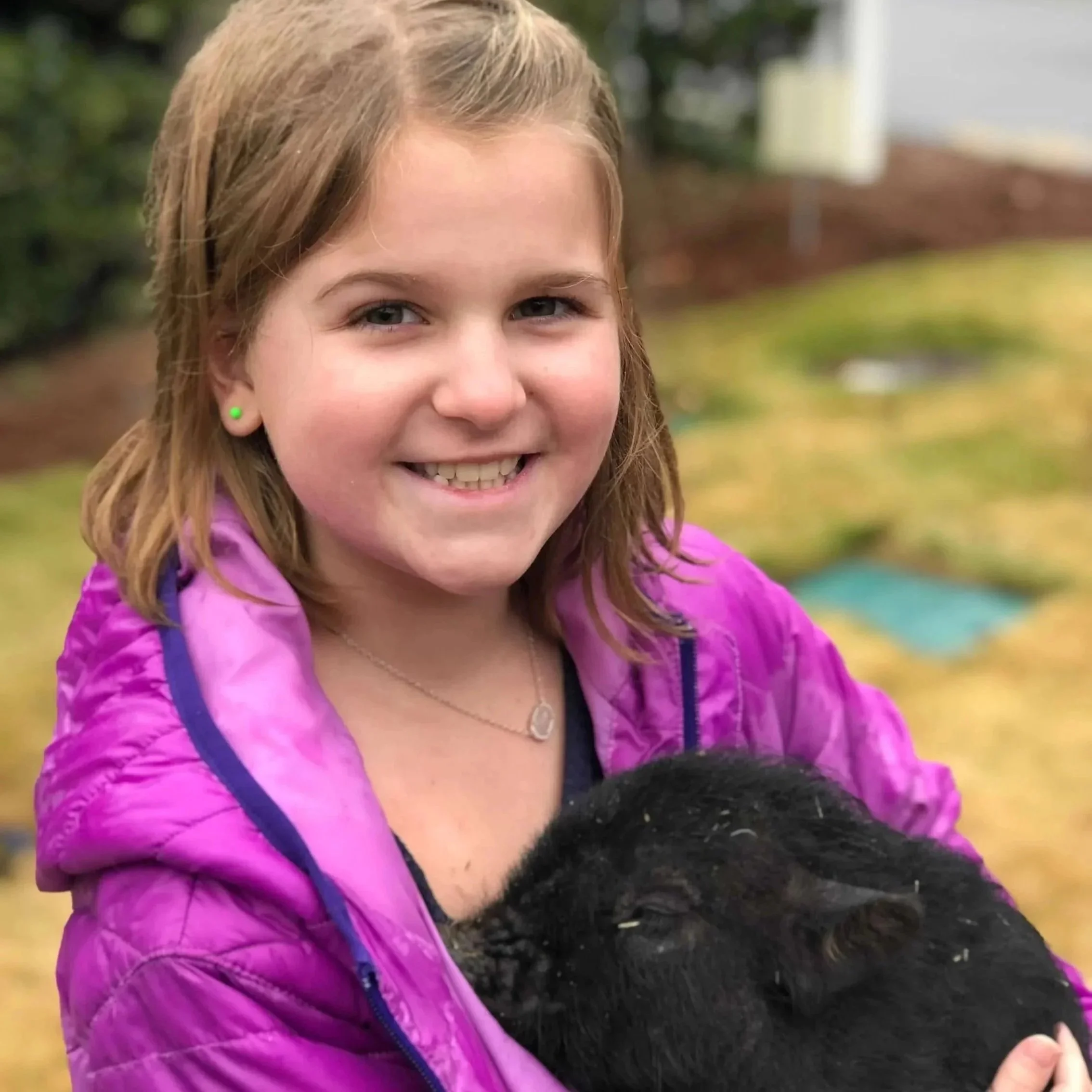 A young girl with light brown hair, blue eyes, and a bright smile, wearing a pink jacket and green earrings, outdoors next to a large black dog with curly fur.