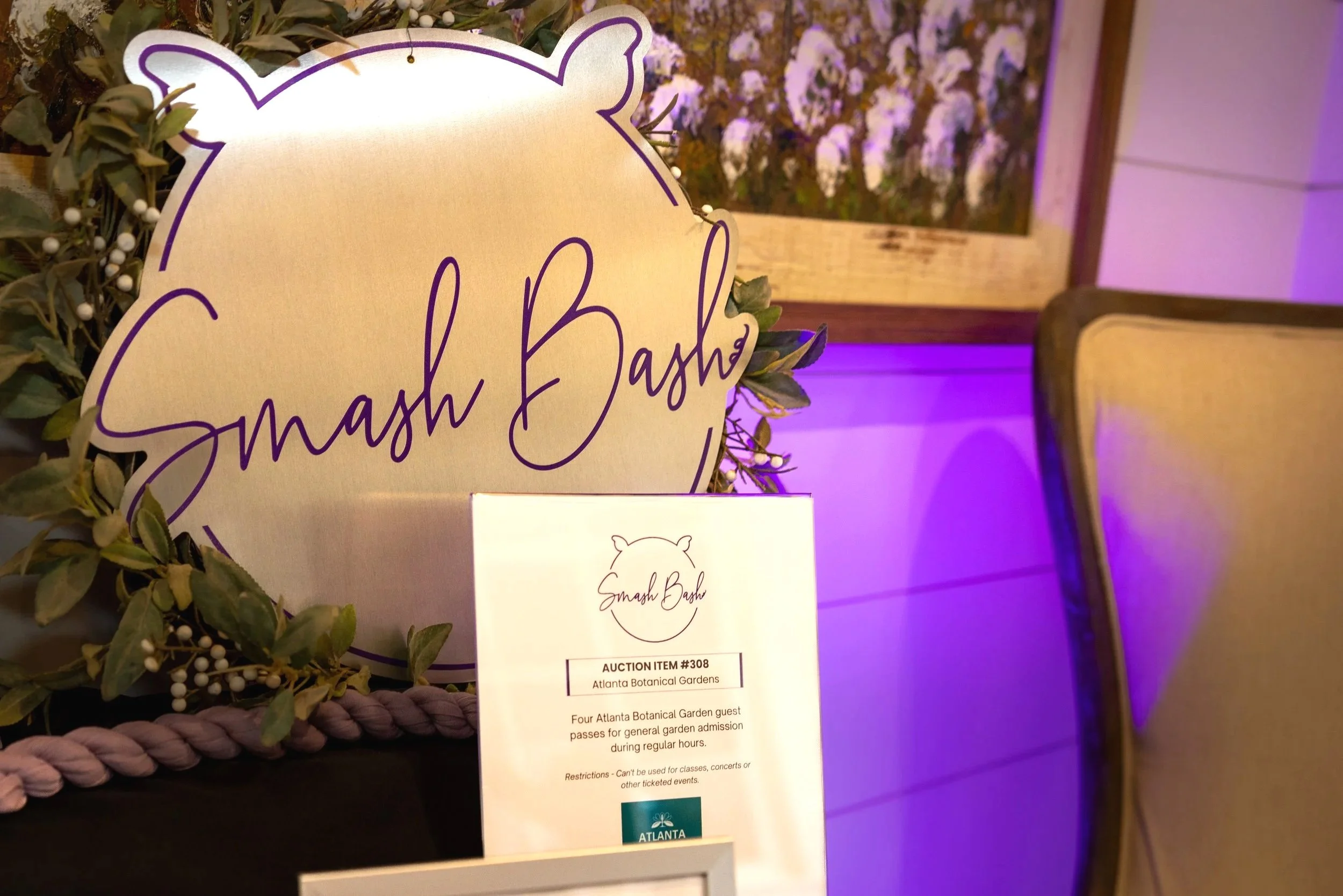 A sign for Smash Bash displayed among green leaves and white berries at the Atlanta Botanical Gardens, with purple lighting in the background.