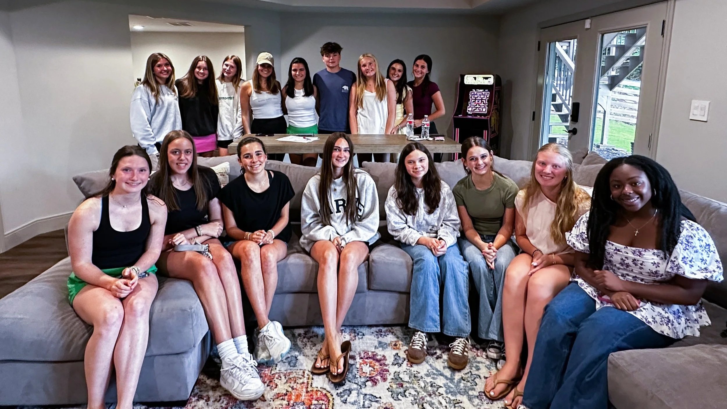 A group of 14 young females and one young male sitting and standing in a living room, with a beige sofa, a rug, a wooden table, and a slot machine in the background.