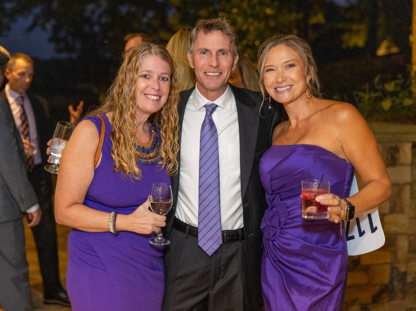Three people dressed in formal attire at an outdoor event, holding drinks and smiling at the camera. Two women in purple dresses and a man in a suit and tie.