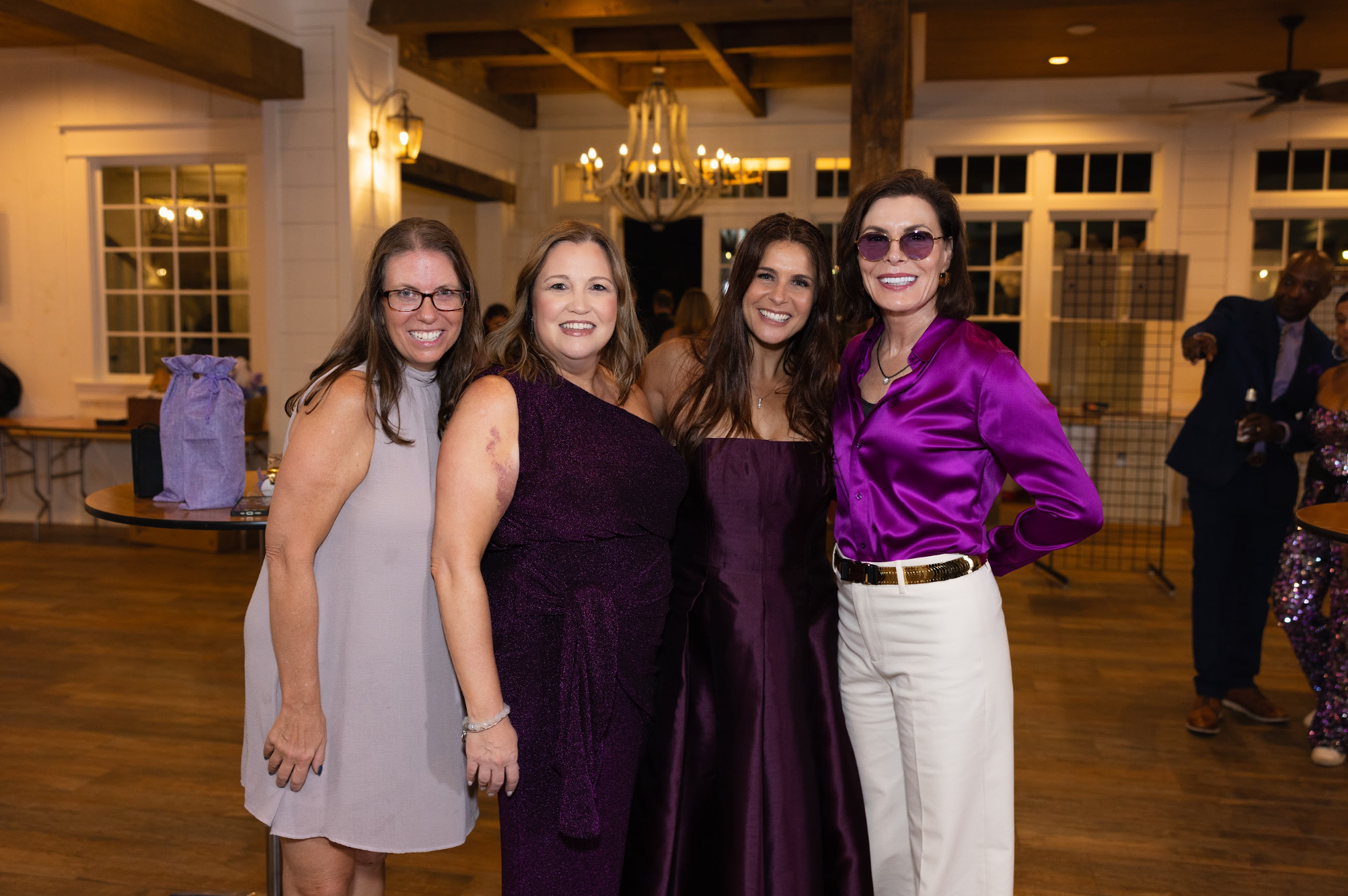 Four women in elegant attire smiling at the camera indoors at a social event, with other guests and warm lighting in the background.