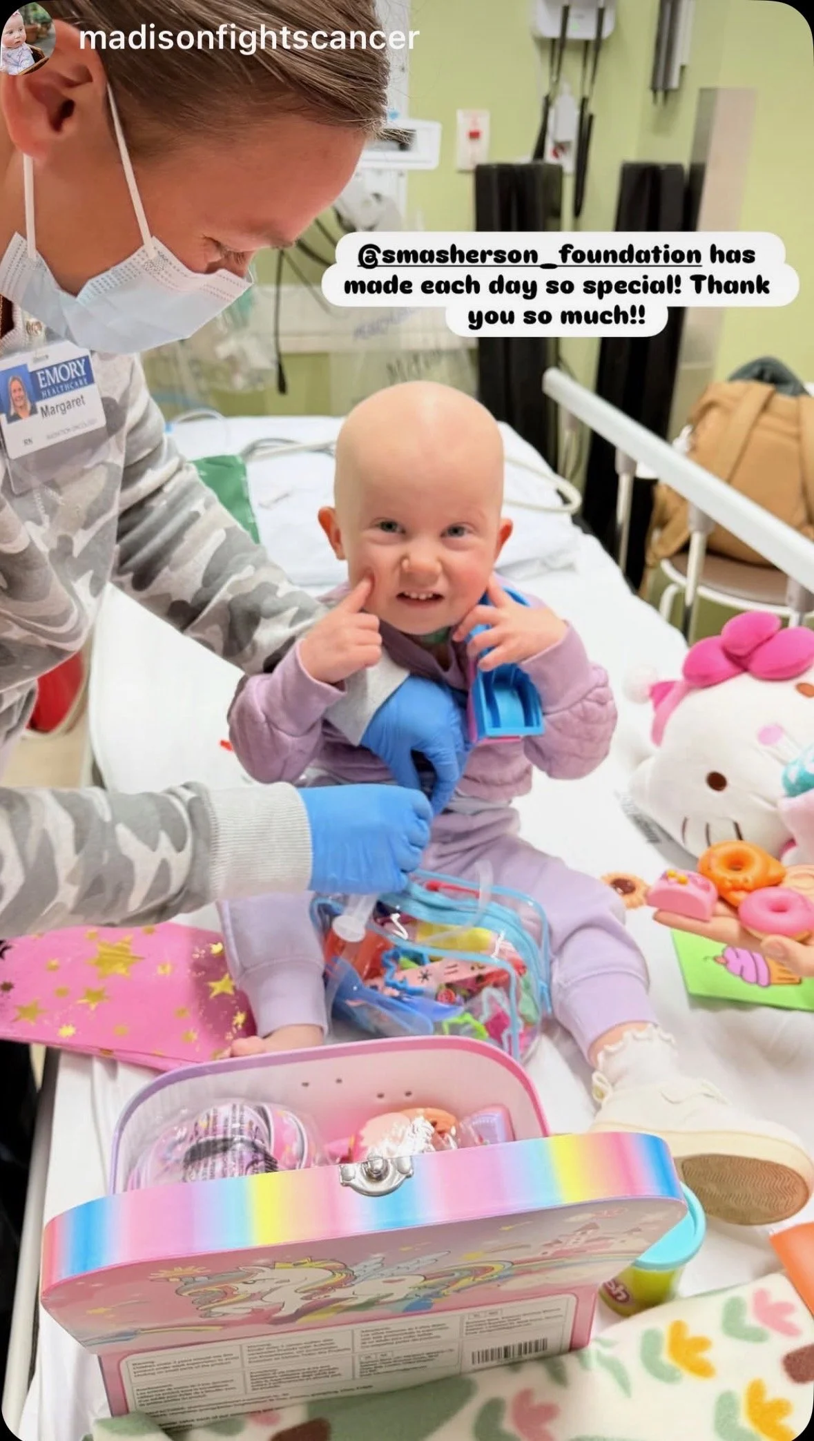 A young child with a shaved head sitting in a hospital bed, smiling and pointing to her cheeks, surrounded by medical equipment and toys. A healthcare worker wearing a mask and gloves is assisting her.