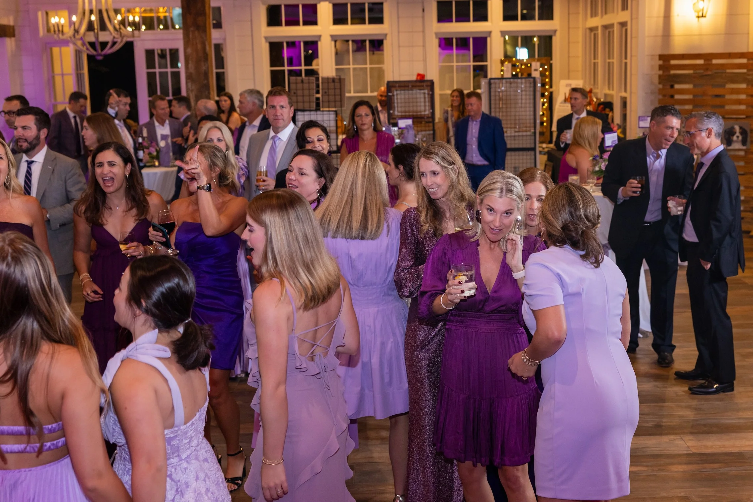 People attending a social event or party in a decorated indoor venue, some holding drinks, with a mix of men and women dressed in formal or semi-formal attire.