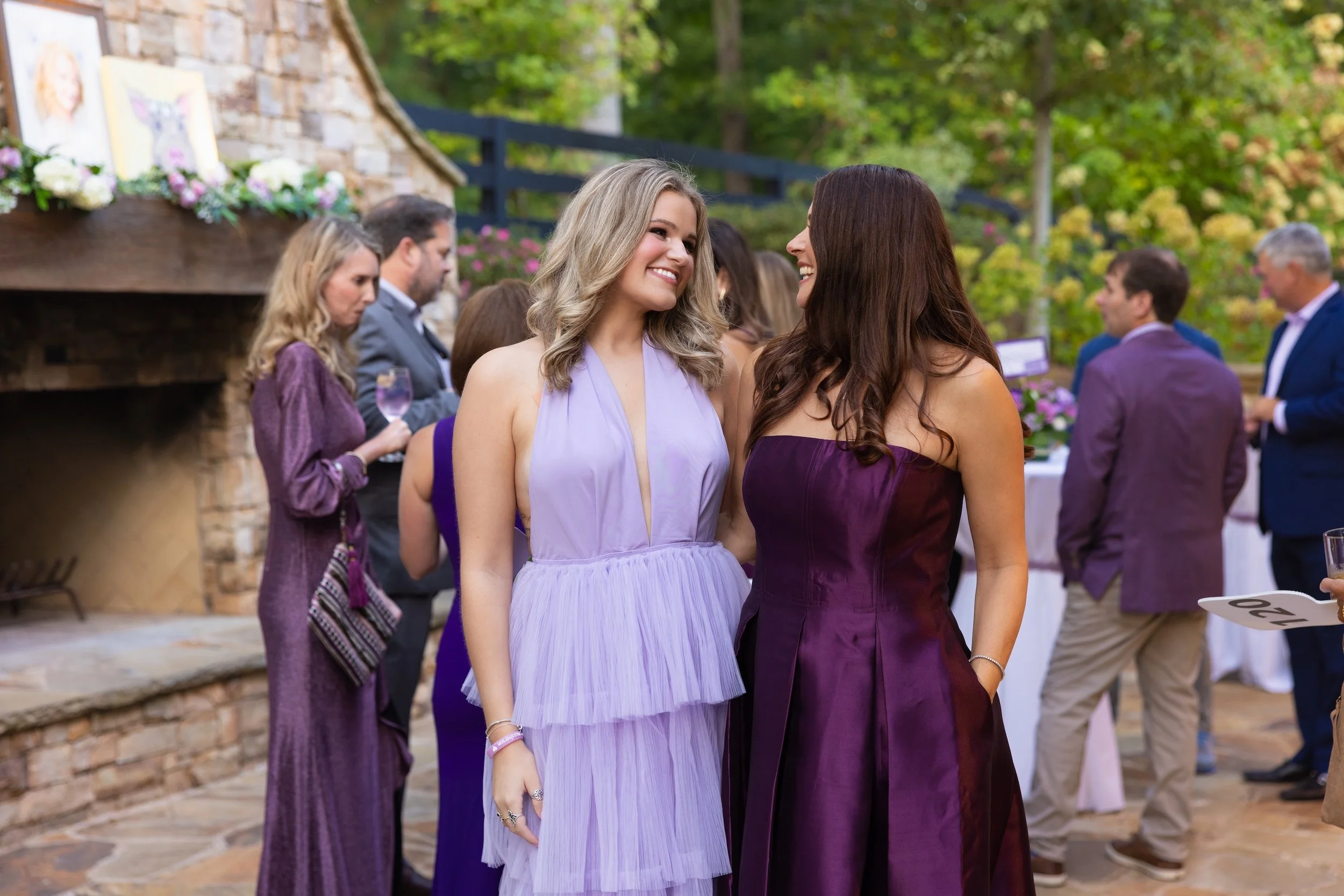 Two women in formal dresses, smiling and conversing at an outdoor gathering with other guests in the background, some holding drinks, surrounded by greenery and a brick structure.
