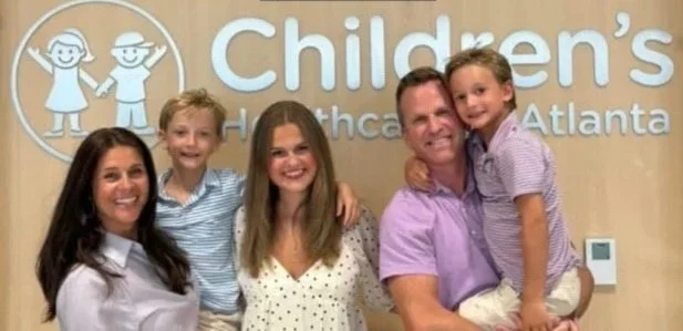 A group of five people, including two children and three adults, smiling and posing in front of a sign that reads 'Children's Healthcare Atlanta.' Two people are holding children, and all are smiling.