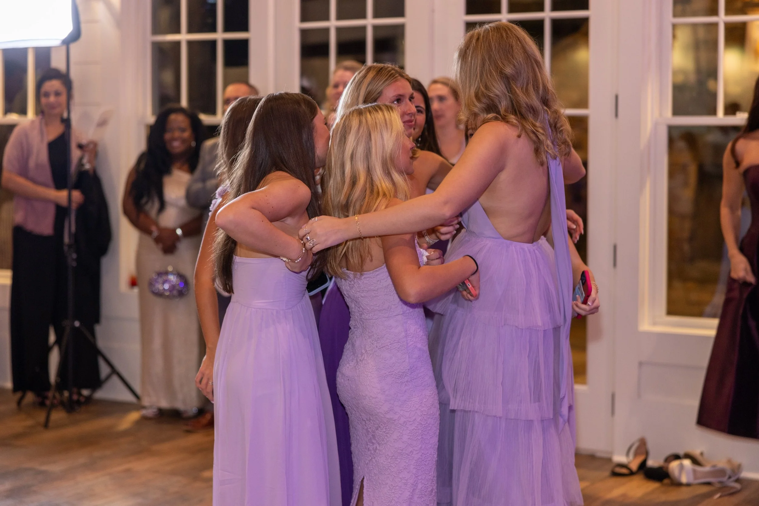 A group of women in matching lavender dresses hugging and celebrating at an indoor event.