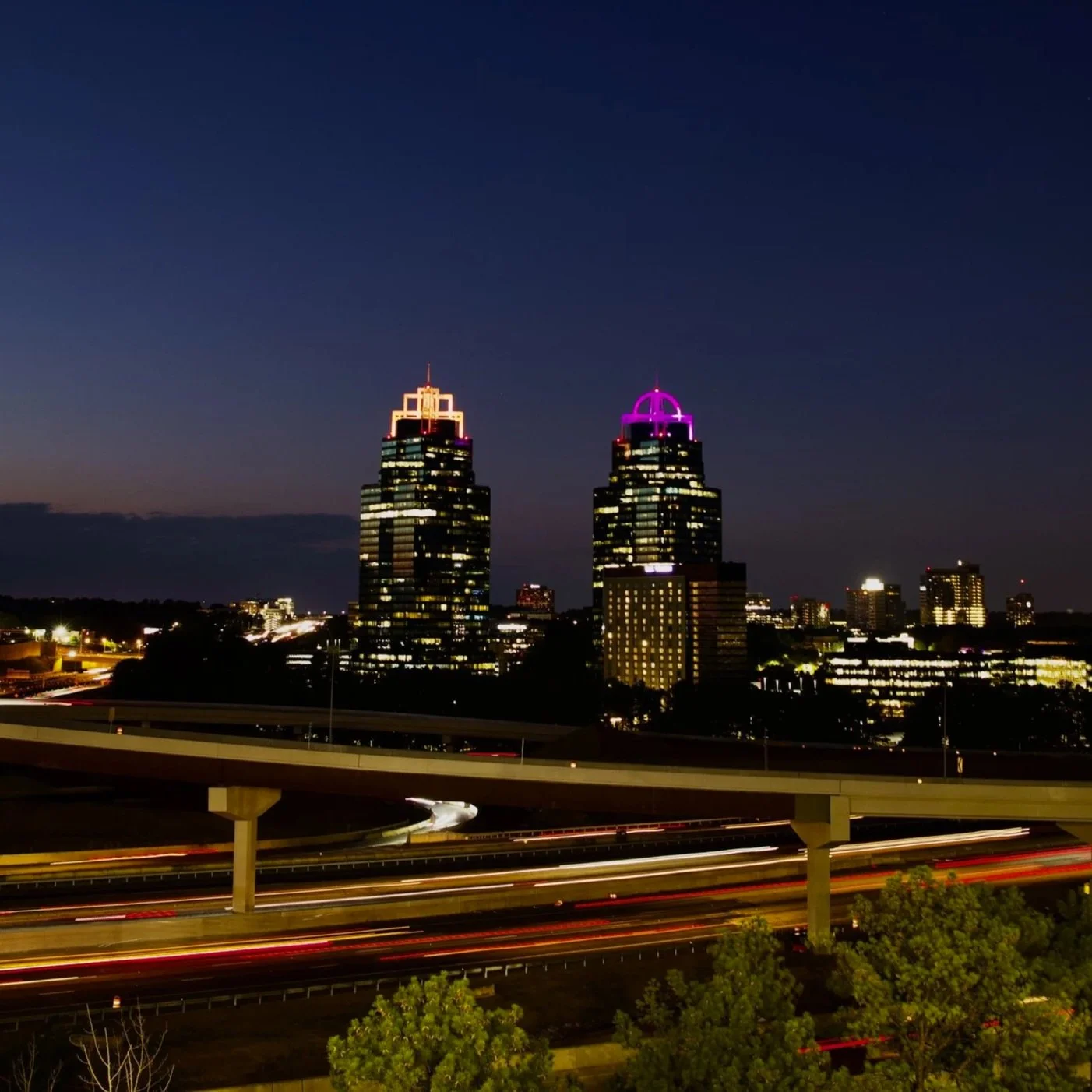 Nighttime cityscape featuring two tall buildings with illuminated tops, pulsating purple and white lights, visible highway with streaks of vehicle lights, and a dark sky in the background.