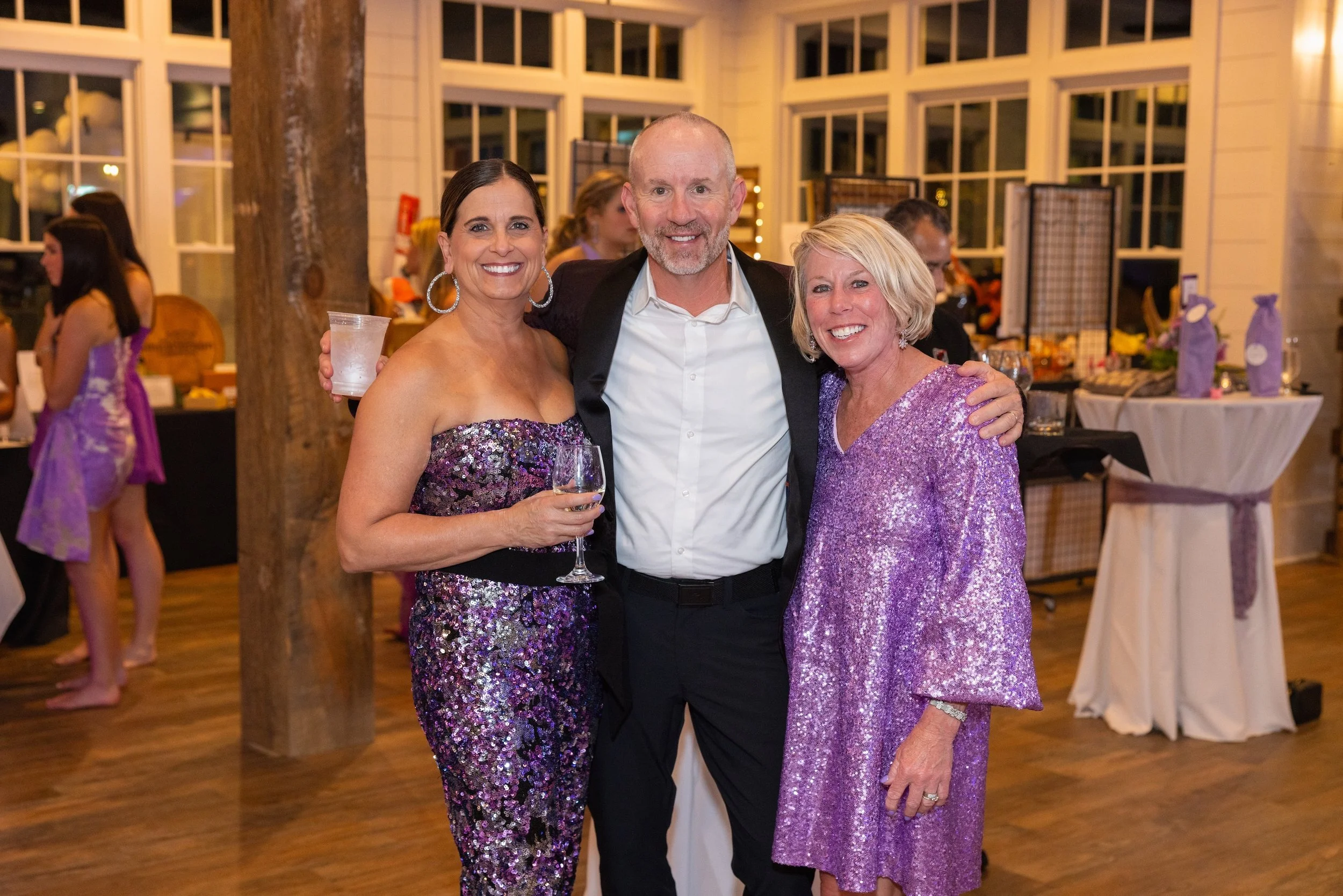 Three people at a social event, two women and one man, smiling and posing for the photo. The women are wearing purple, sparkly dresses and the man is dressed in a white shirt with a black jacket. The background shows other guests and a decorated indo