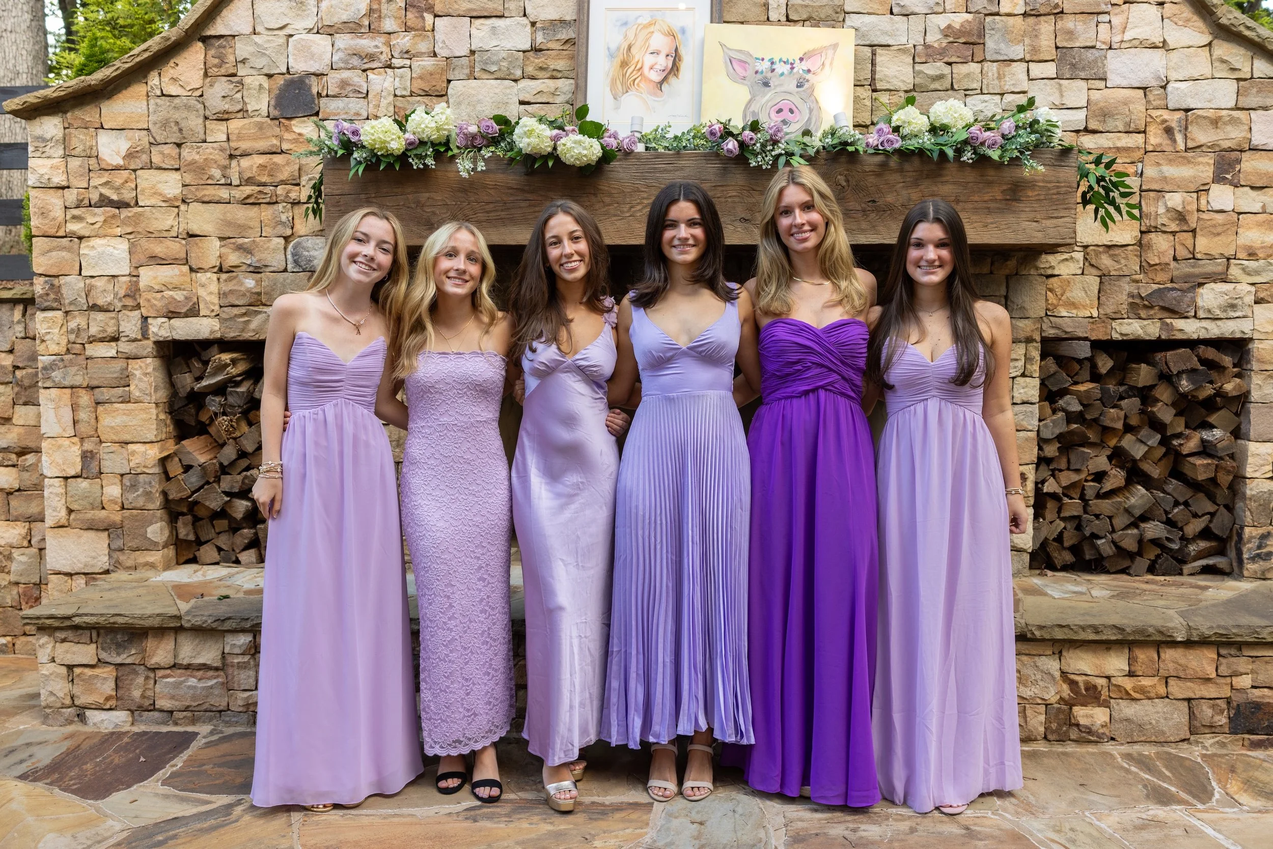 Six women in purple dresses standing in front of a brick fireplace with artwork and flowers on mantel.