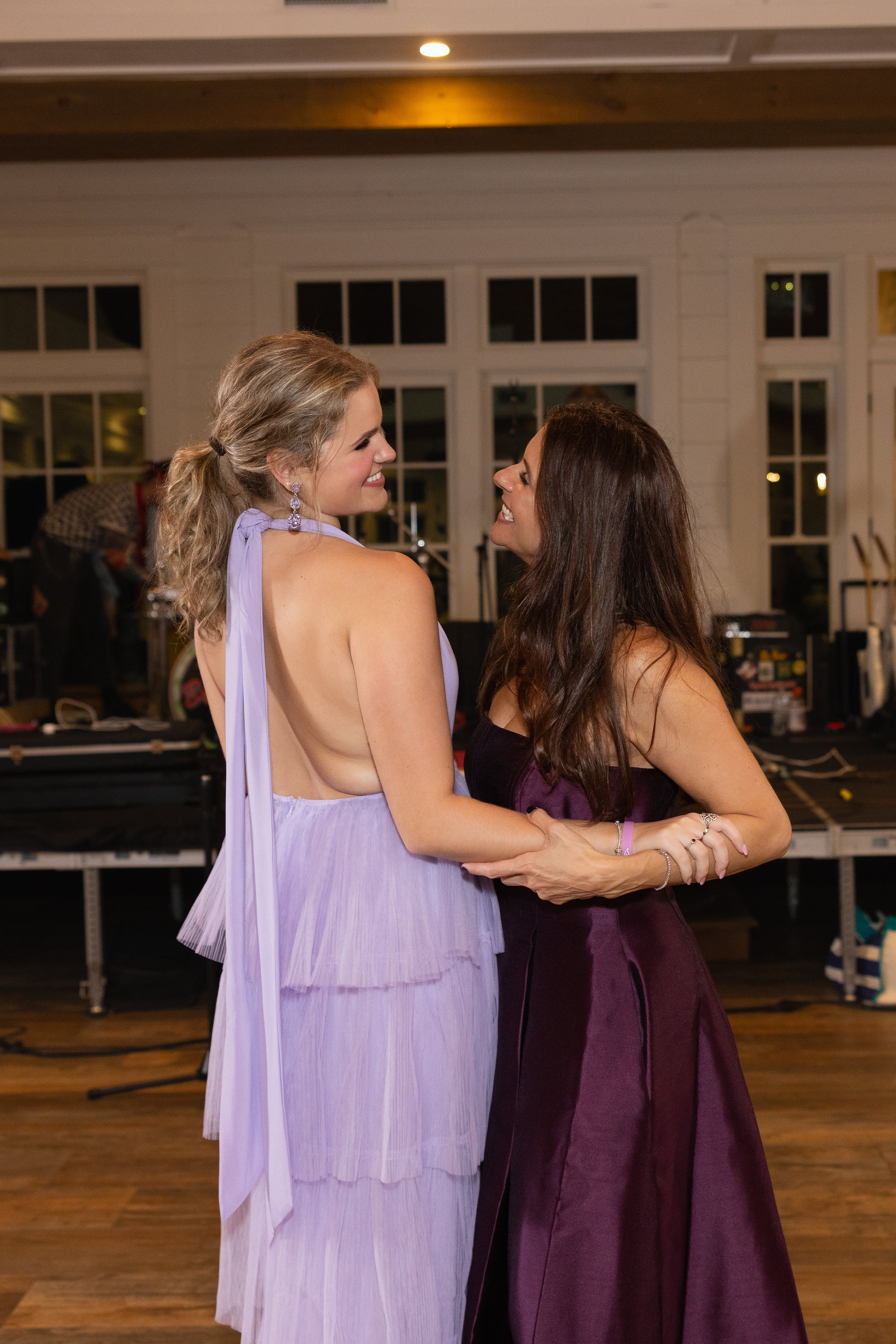 Two women smiling and dancing closely indoors, with large windows and wooden floors, in an elegant setting.