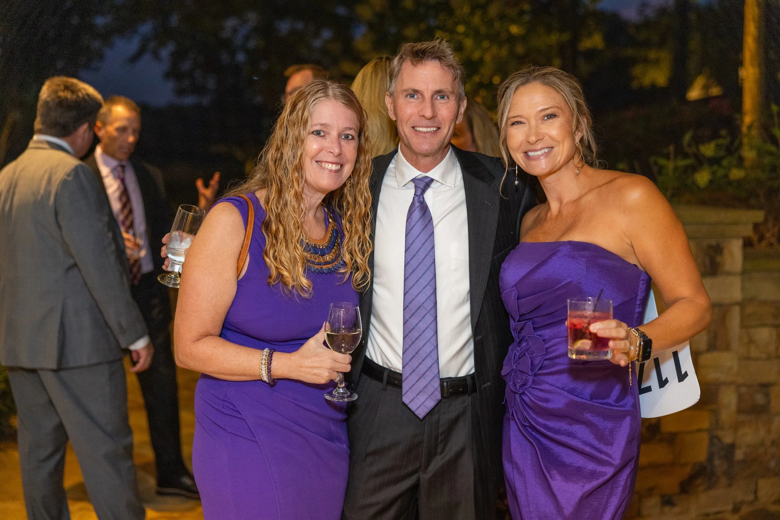 Three adults at an evening outdoor event, dressed in formal attire, holding drinks and smiling at the camera. Two women in purple dresses and a man in a suit with a purple tie.