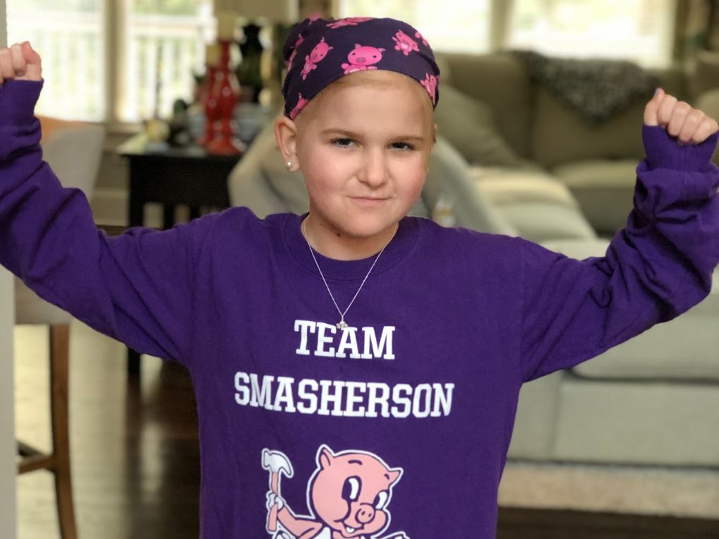 A young girl wearing a purple sweatshirt with the text "Team Smasherson" and a cartoon pig graphic, flexing her arms and smiling.