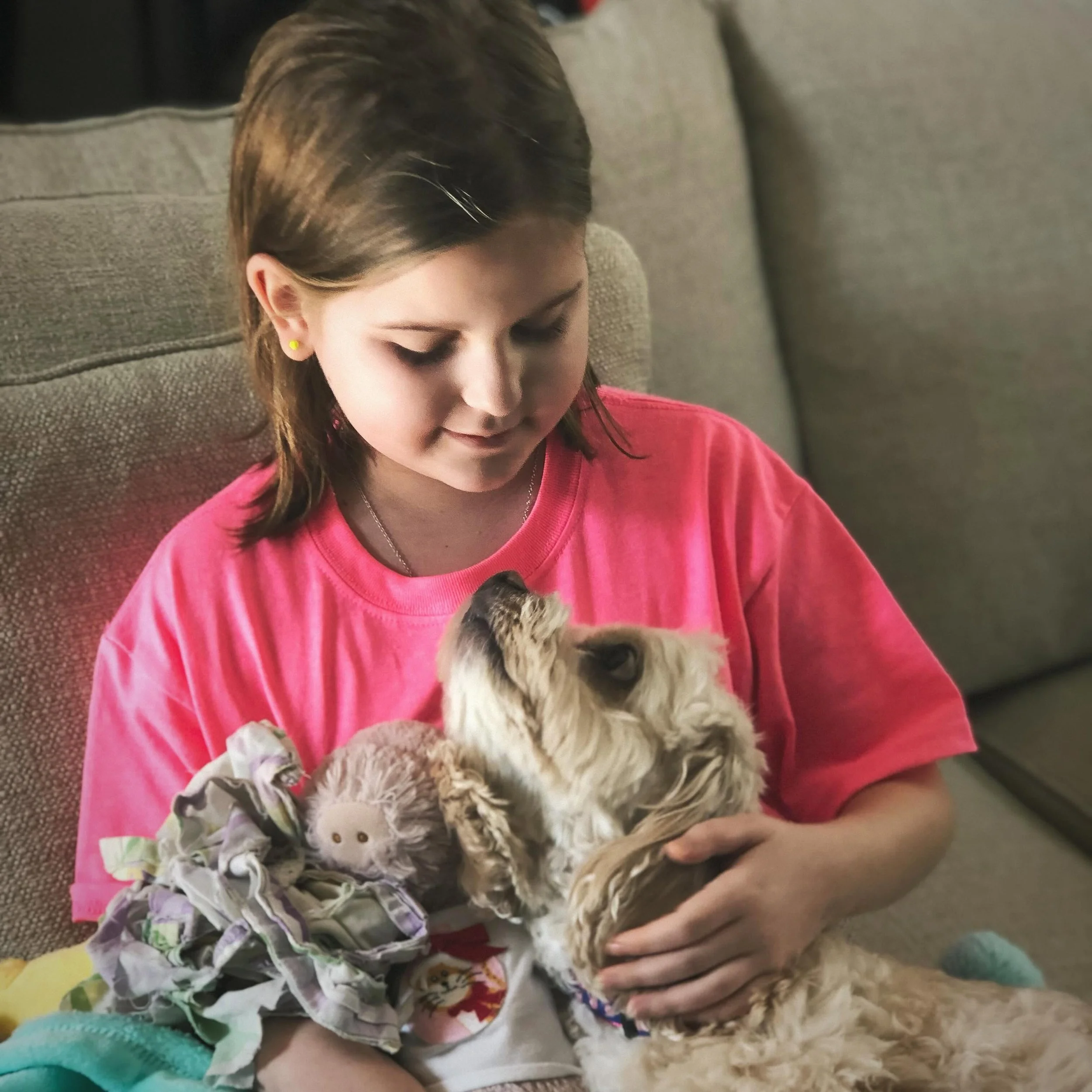A young girl with brown hair and yellow earrings wearing a pink shirt sitting on a couch, holding and looking at a small puppy with curly fur.