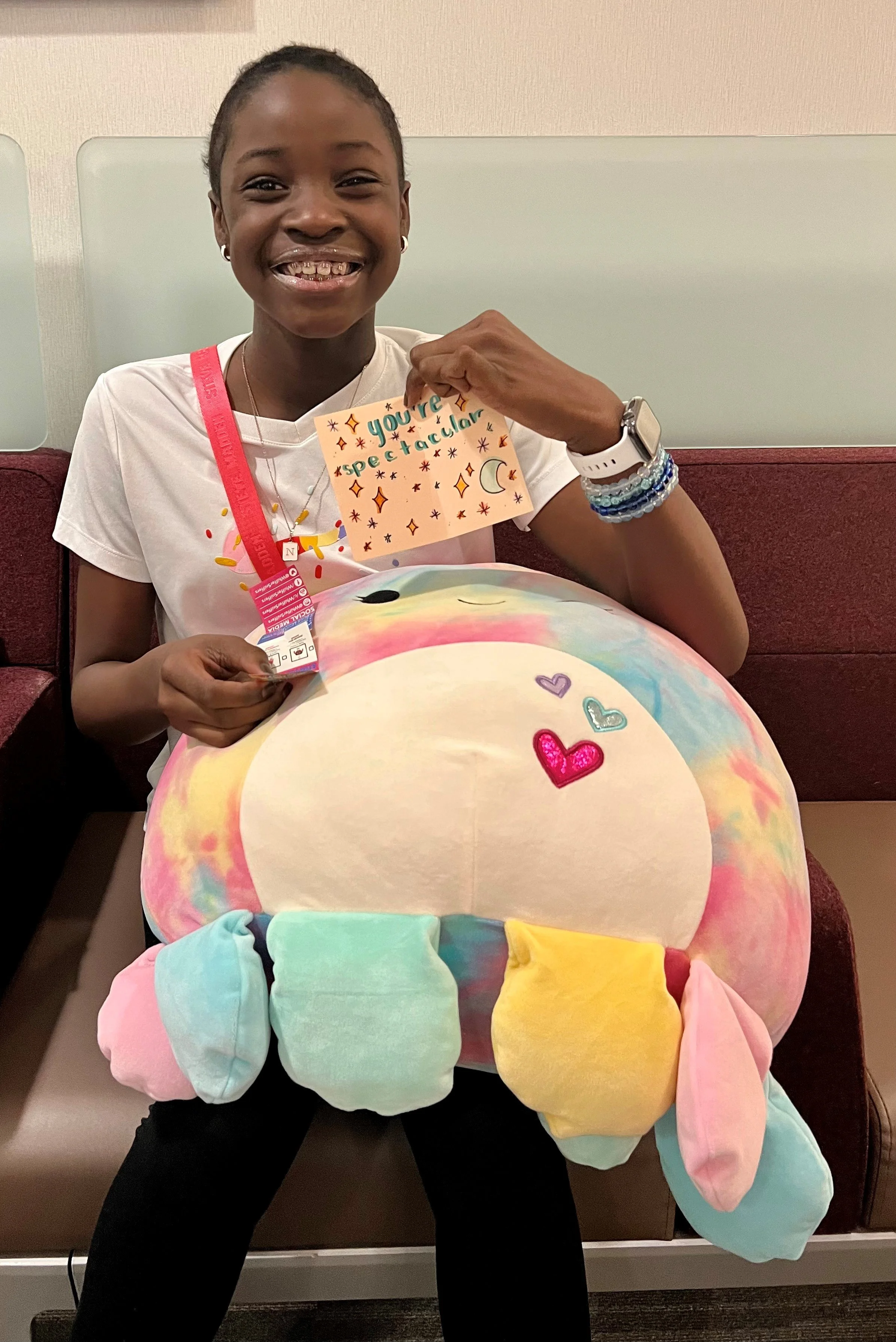 A young girl with short hair and a big smile, holding a colorful plush unicorn pillow, sitting on a bench at a hospital, wearing a white shirt, a lanyard, and a watch.