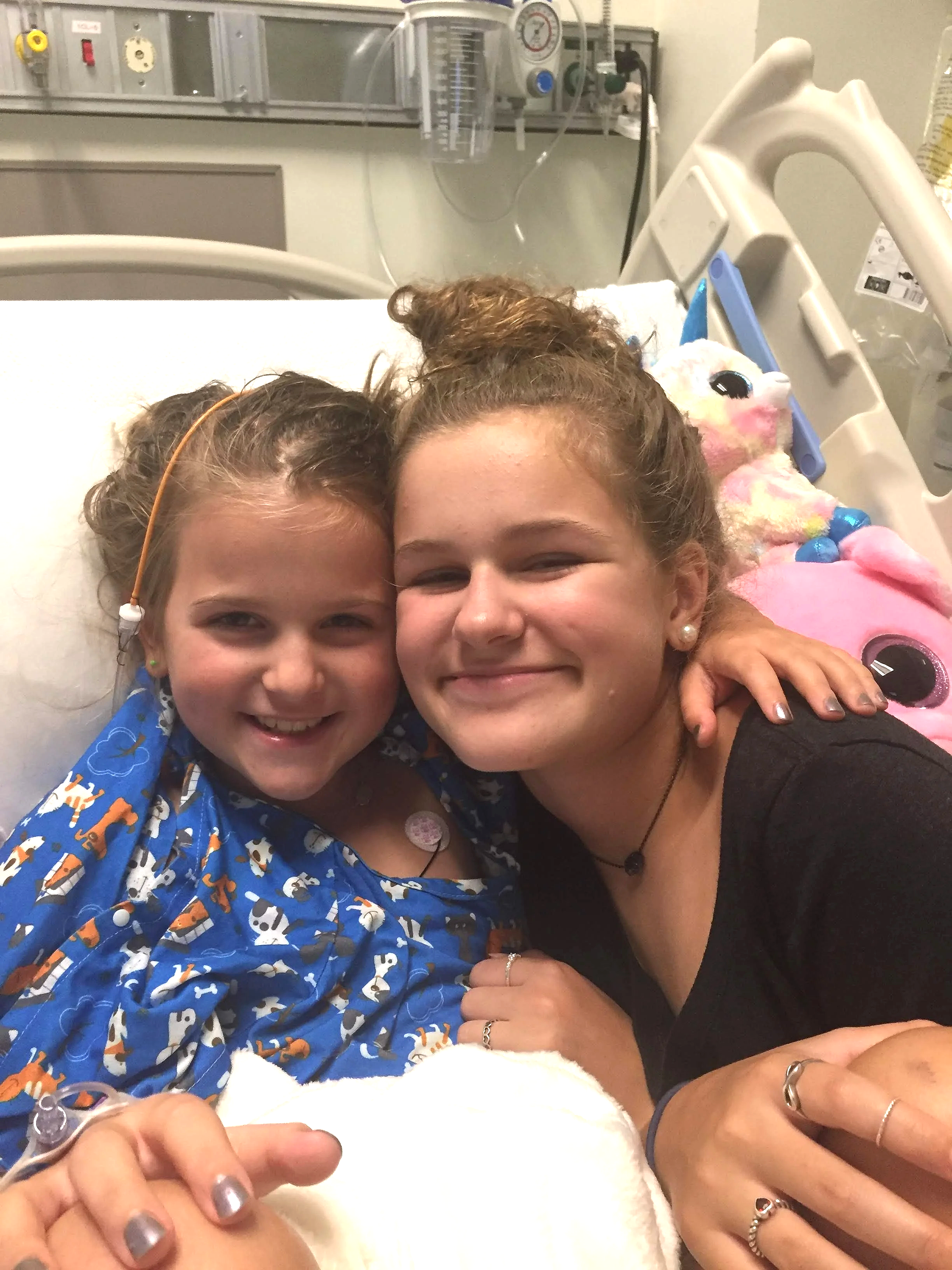 Two young girls lying in a hospital bed, smiling and holding hands. One girl is wearing a blue shirt with a dog pattern, the other in a black shirt. They are in a medical setting with tubing, medical devices, and a plush toy nearby.