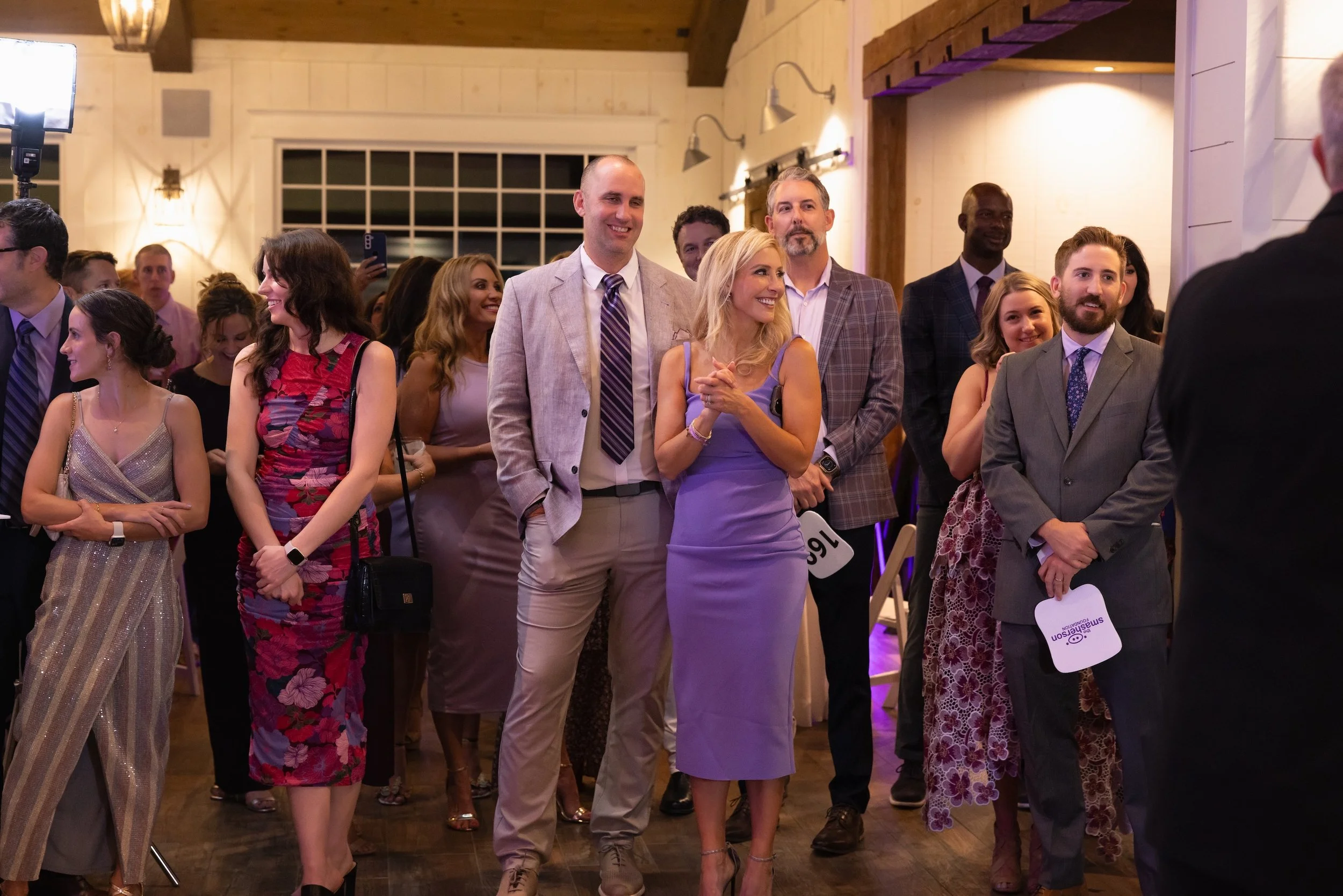A group of people dressed in formal attire attending a wedding or celebration event, standing indoors with wooden walls and warm lighting.