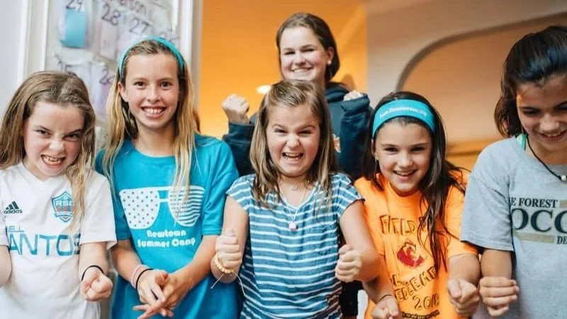 Group of smiling girls at a summer camp, some making fists, in a cheerful indoor setting.