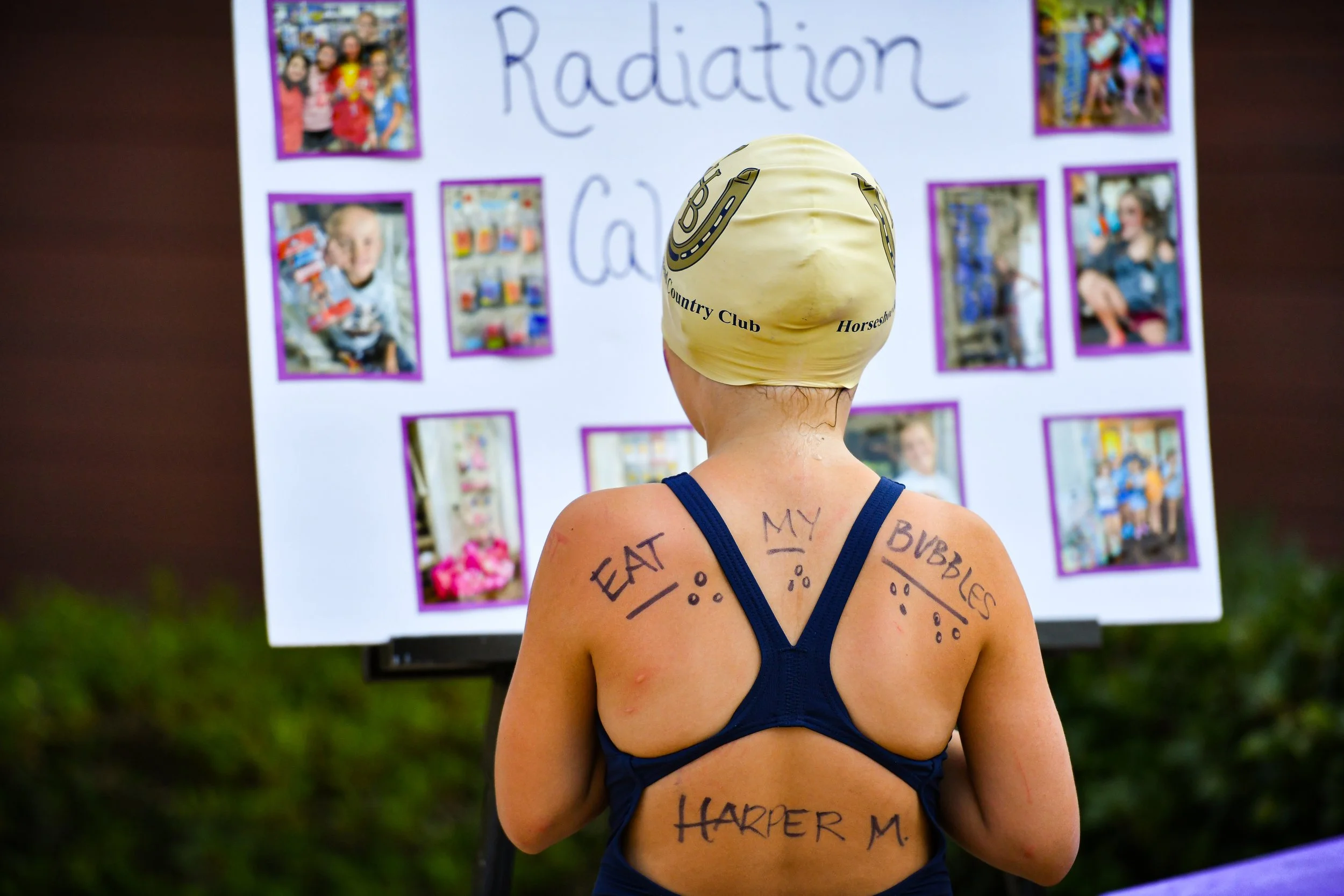 A person wearing a swim cap with a horseshoe symbol and the words 'Country Club' on it, standing in front of a display board with photos and the word 'Radiation' written at the top. The person's back is covered with handwritten messages.